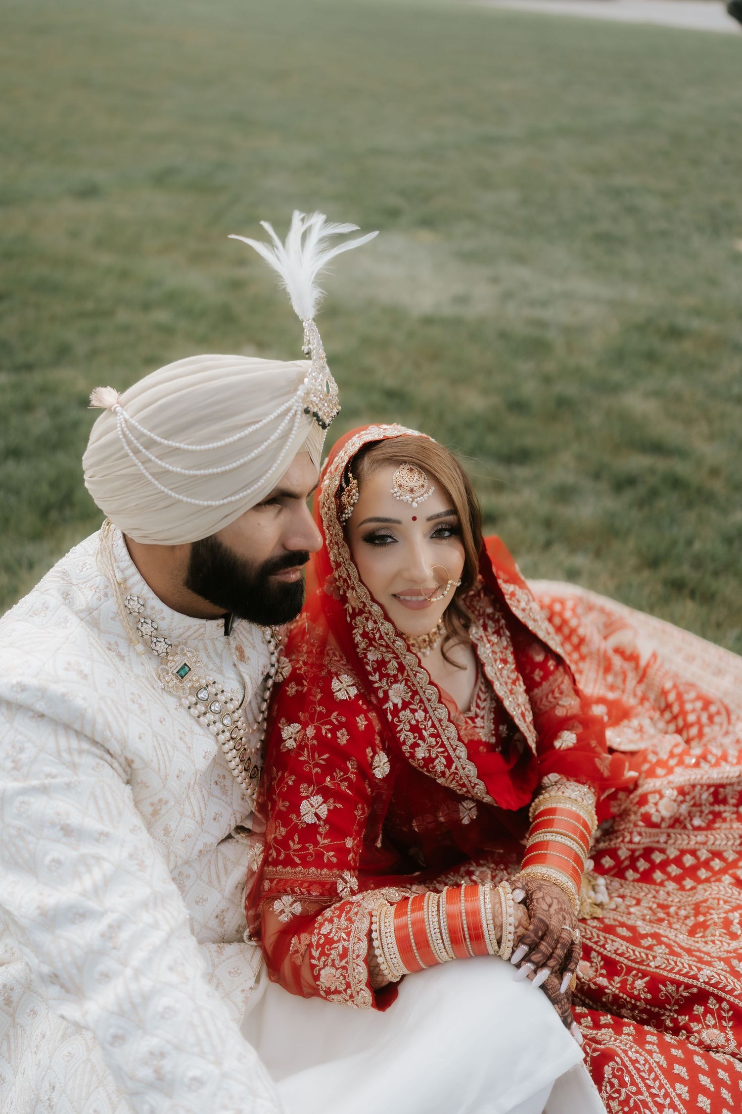 Sikh bride in red lehenga and groom in white sherwani and turban sitting together on grass at their wedding.