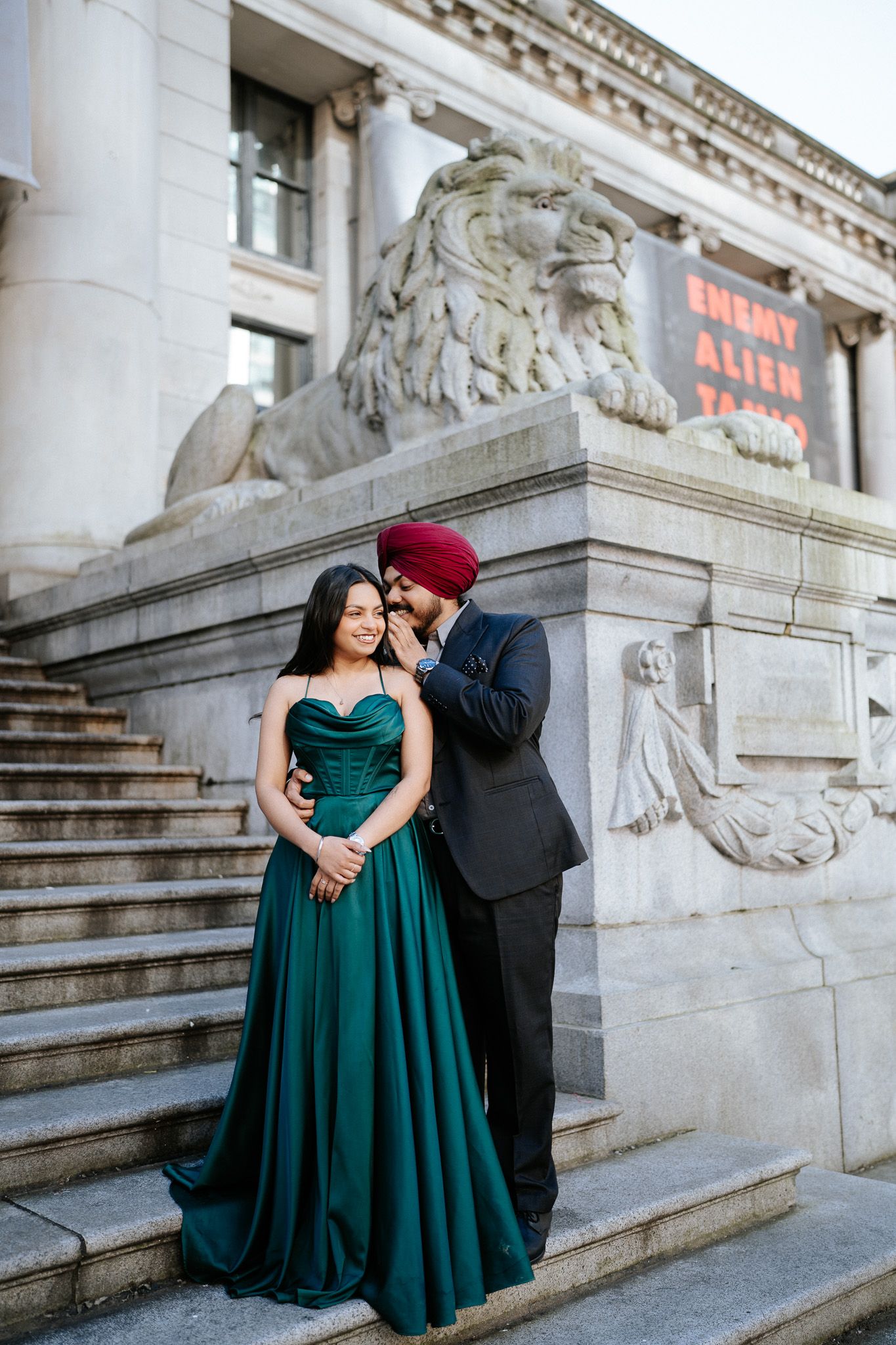 A couple in formal attire poses on ornate stone steps, with the person in a green gown seated on the steps.