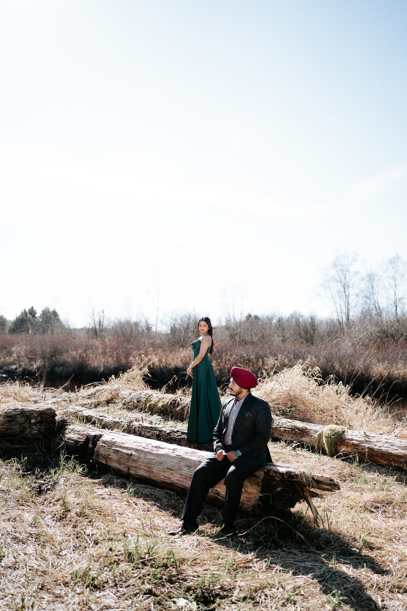 A couple sits together on a fallen log in a winter field during a romantic outdoor photoshoot.
