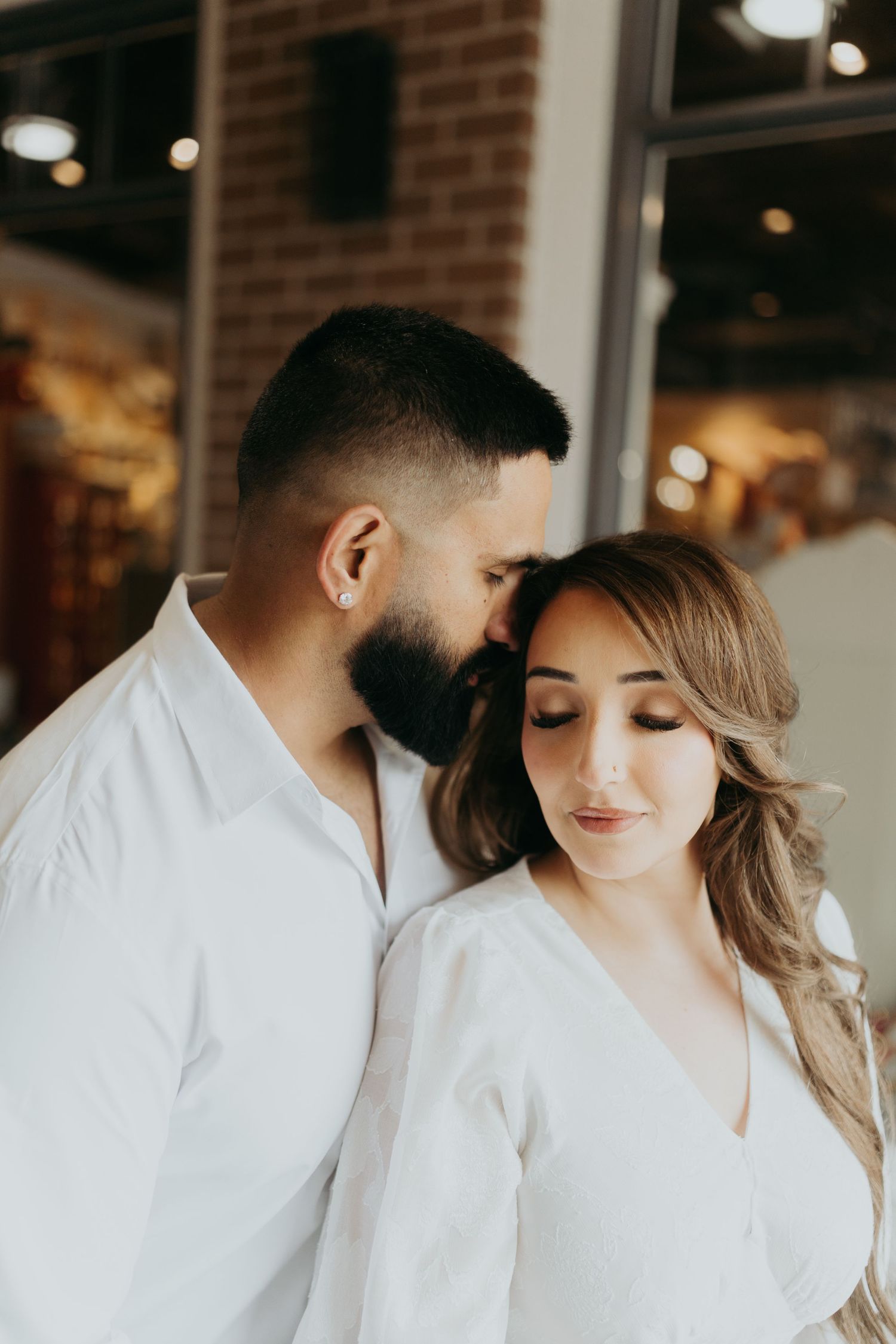 Romantic couple in white outfits share intimate moments in an indoor setting with brick walls and soft lighting.