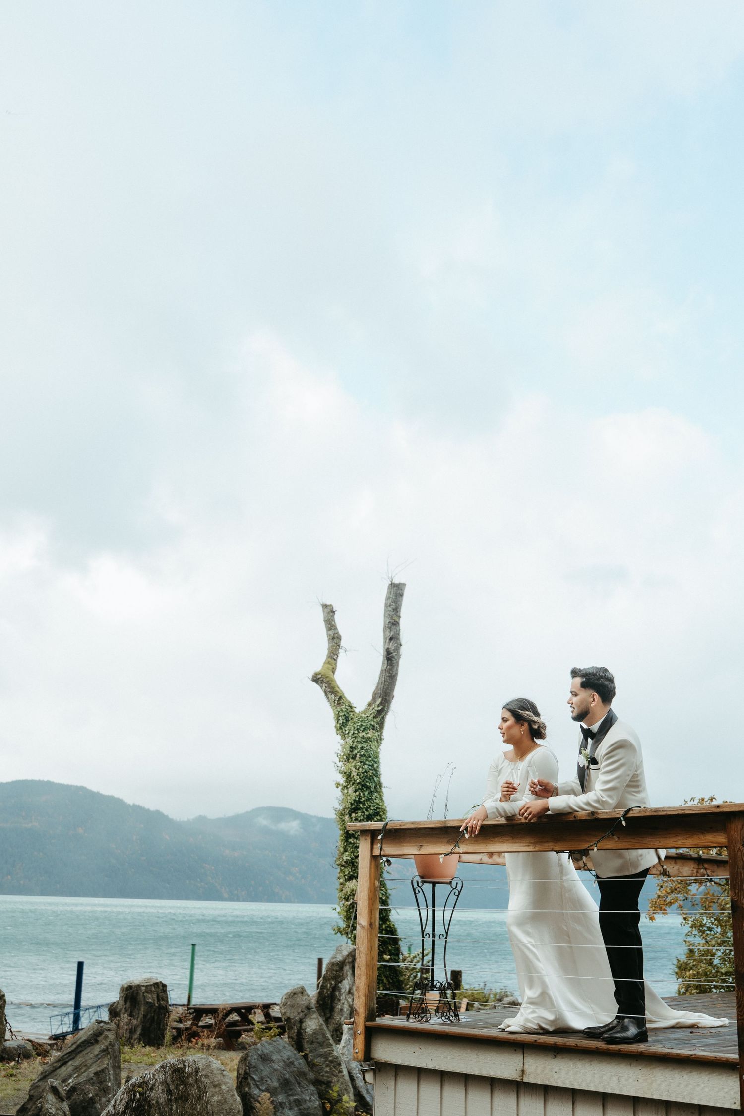 A couple sits on a rustic wooden deck overlooking a misty lake and mountains in a romantic outdoor setting.