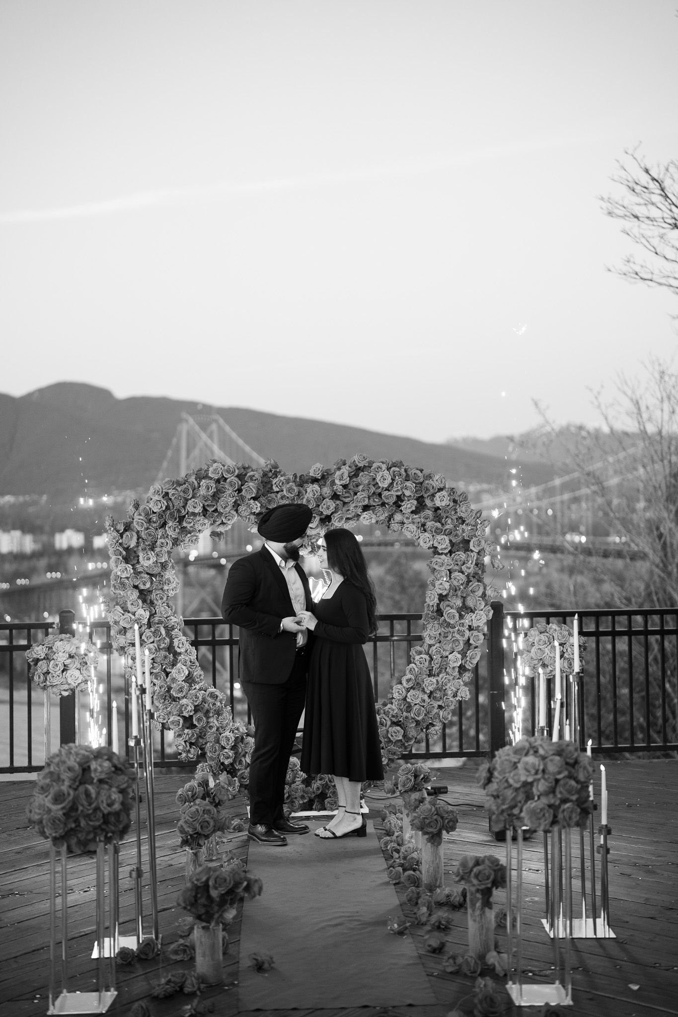 Two figures in dark clothing stand on an outdoor terrace overlooking a scenic mountain landscape in black and white.