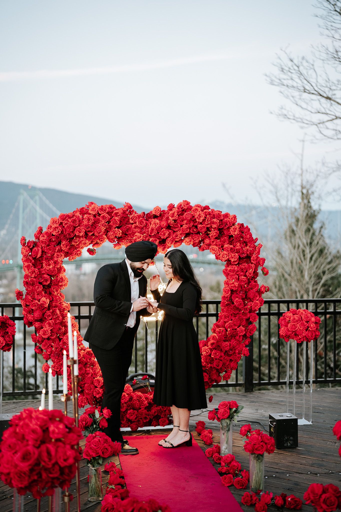 A romantic proposal setup with a bright red floral arch against a city bridge backdrop.
