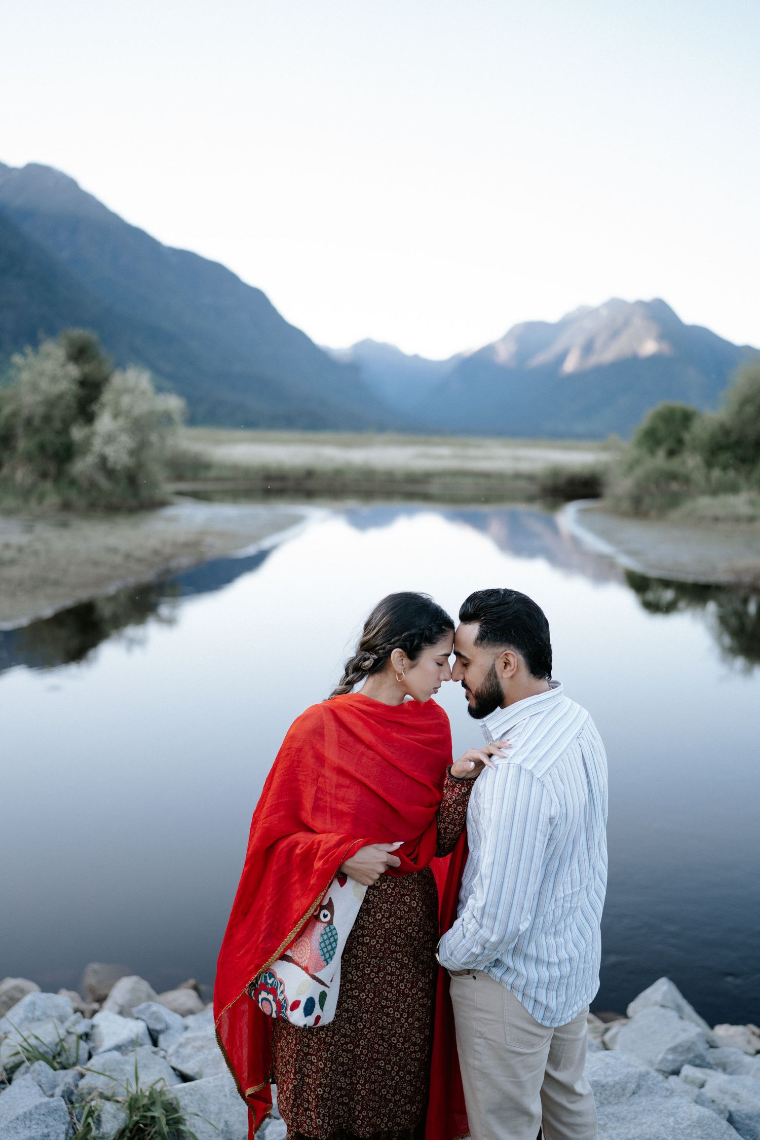 A couple shares an intimate moment by a serene mountain lake at dusk, with the woman wearing a striking red shawl.