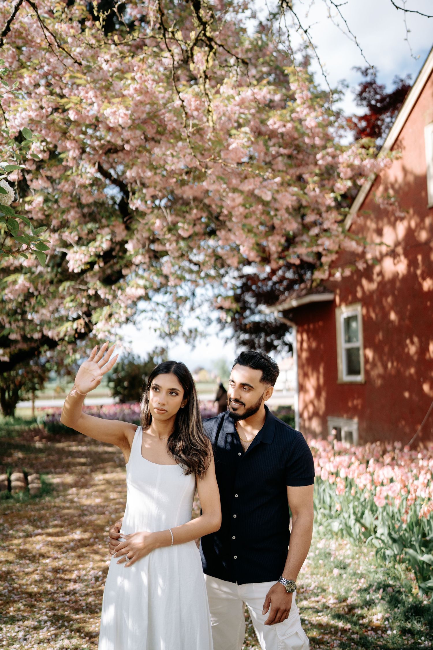 A couple poses together under blooming cherry blossom trees near a red brick building in springtime.