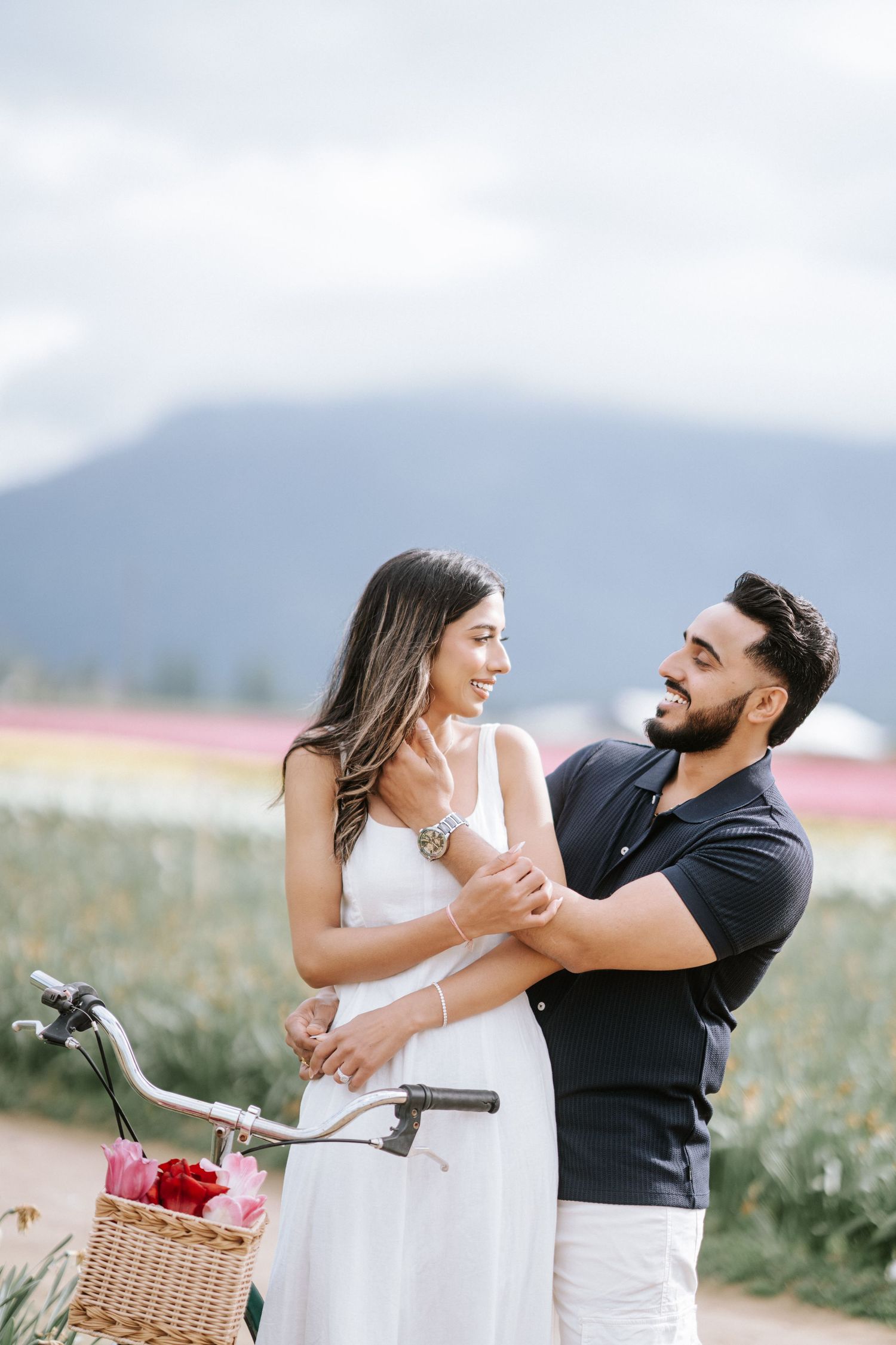 A couple shares a romantic moment in a field with mountains in the background and a bicycle basket visible.