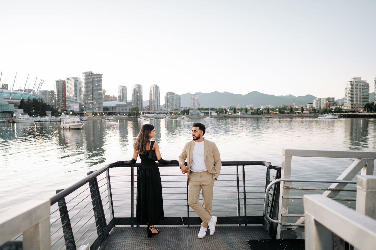 Two people stand on a boat deck overlooking a cityscape with high-rise buildings and a bridge reflected in calm waters.