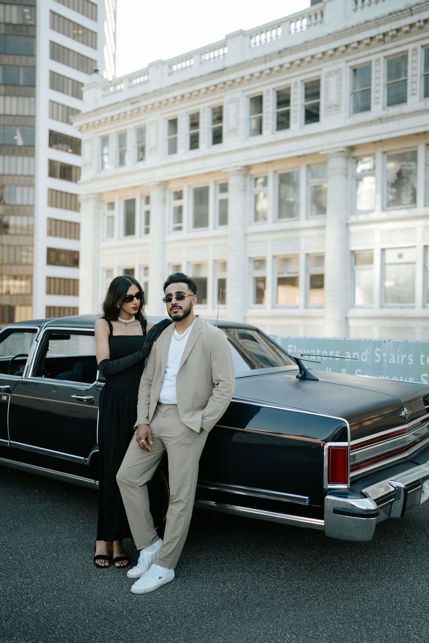 A stylish couple poses with a vintage black Cadillac limousine in an urban setting.