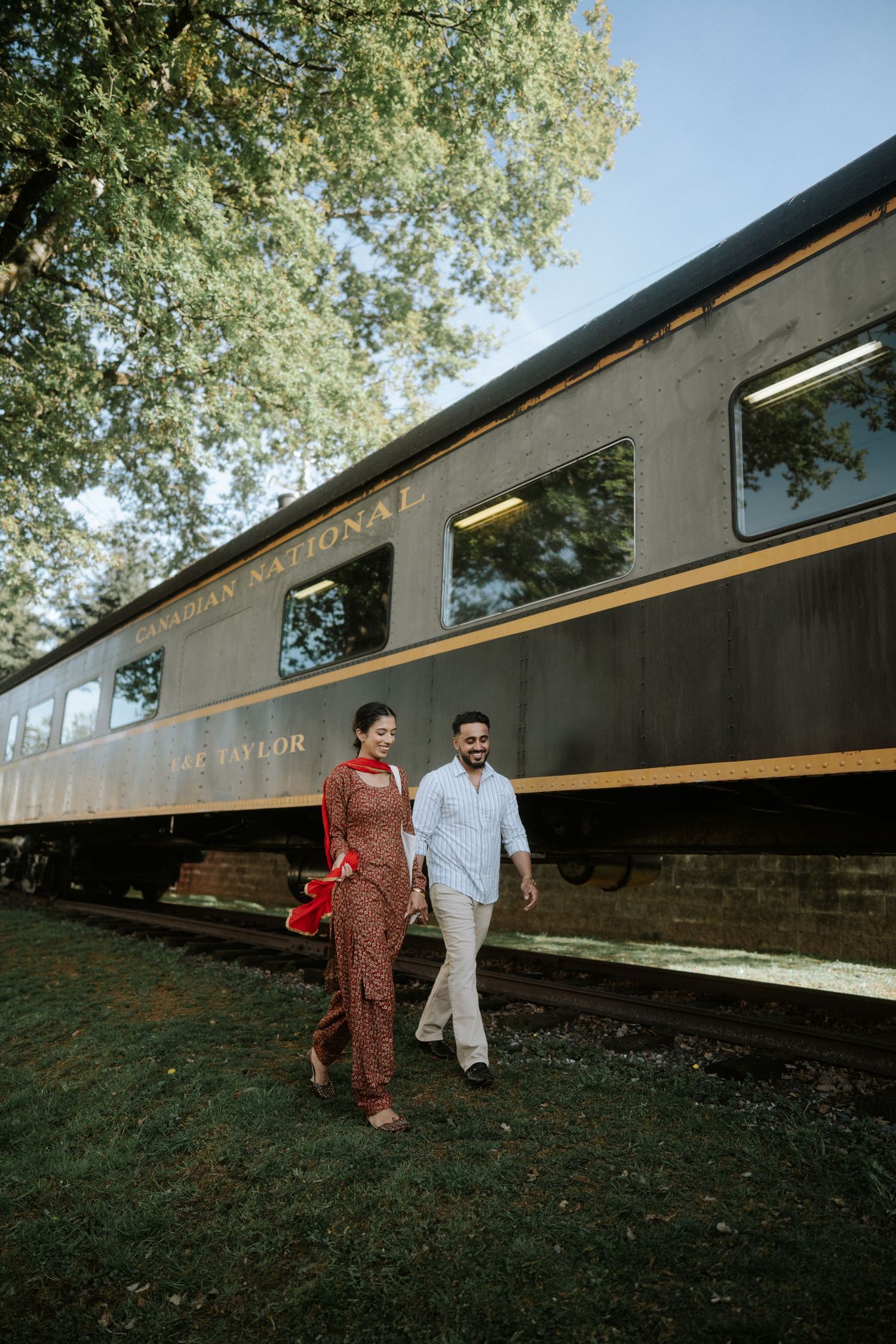 Two people walk along a dark wall with trees visible above during an outdoor photo session.