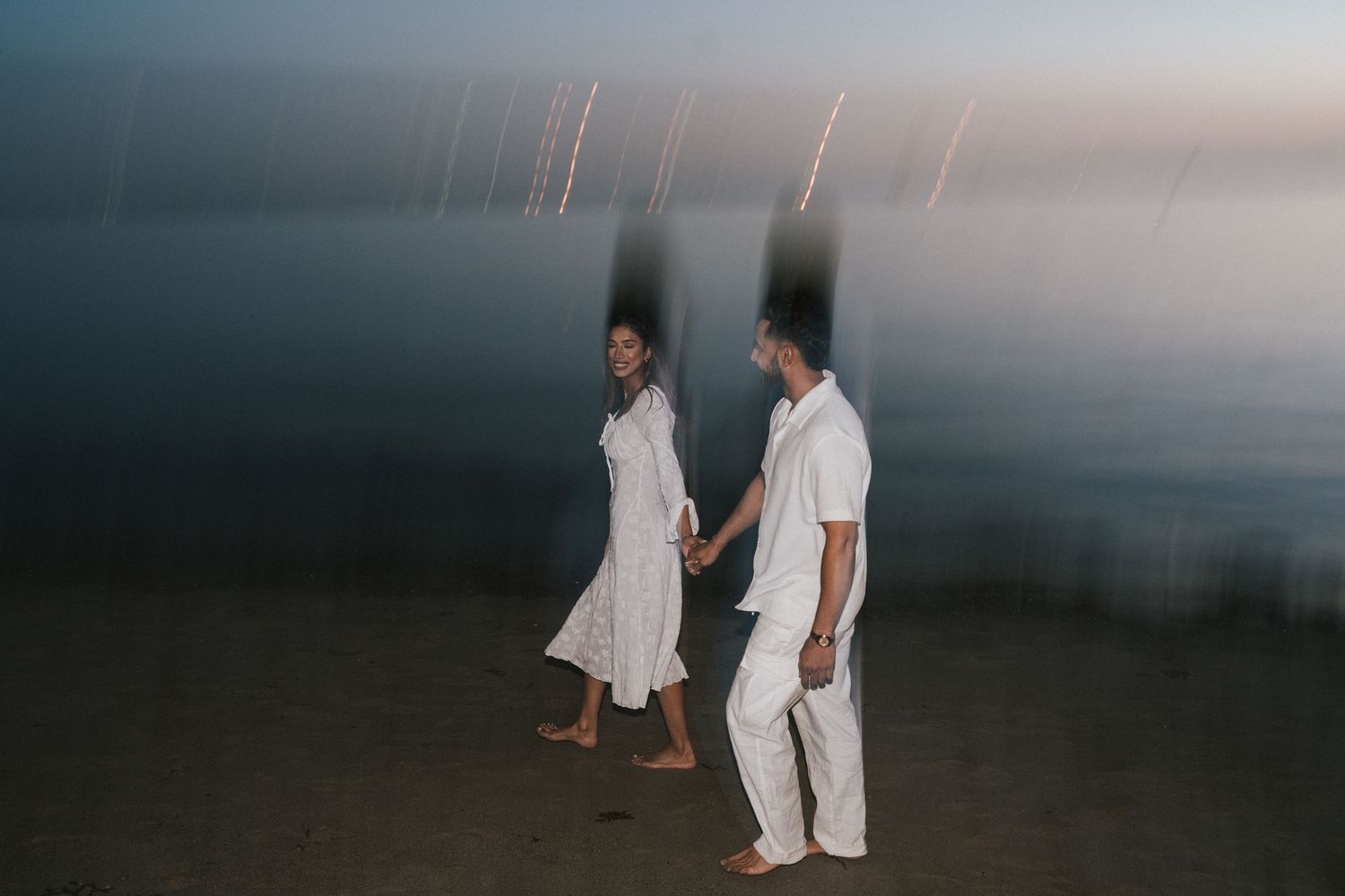 A couple in white outfits walks along a beach at dusk with a moody, dark sky overhead.