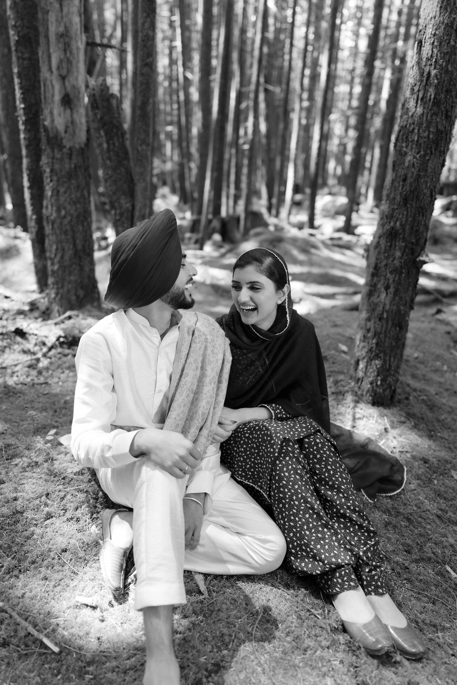 Black and white photo of laughing Sikh couple sitting together on forest ground, man in turban and woman in traditional attire.