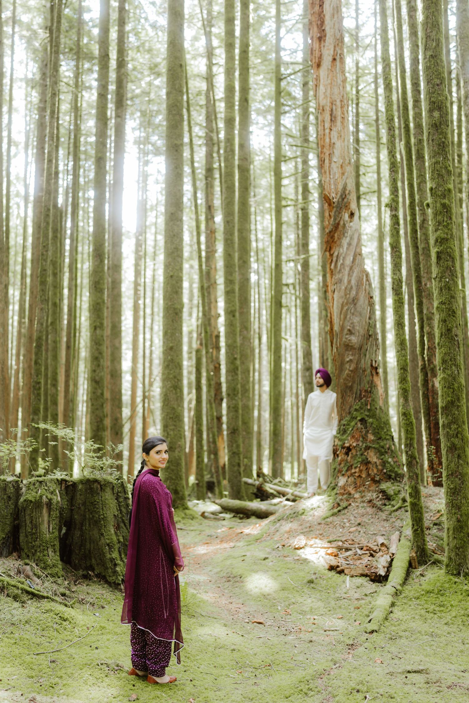 Couple in traditional South Asian attire standing among tall forest trees, woman in burgundy salwar and man in white kurta.