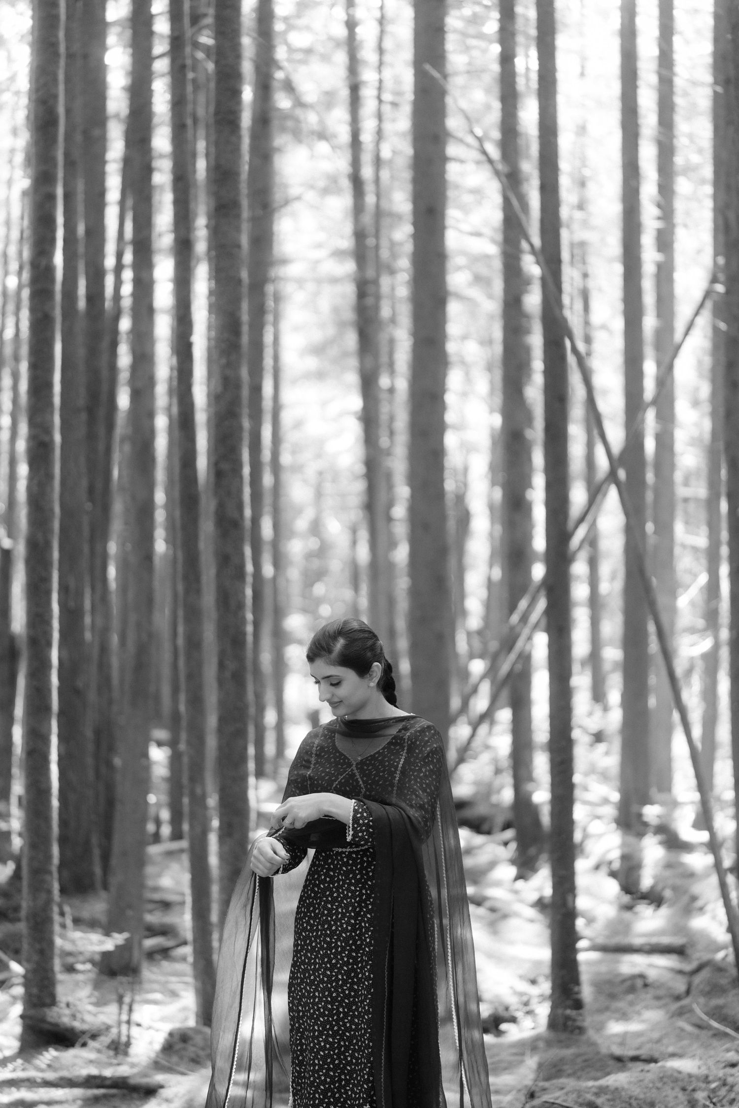 Black and white portrait of a woman in traditional Indian attire standing in a pine forest.