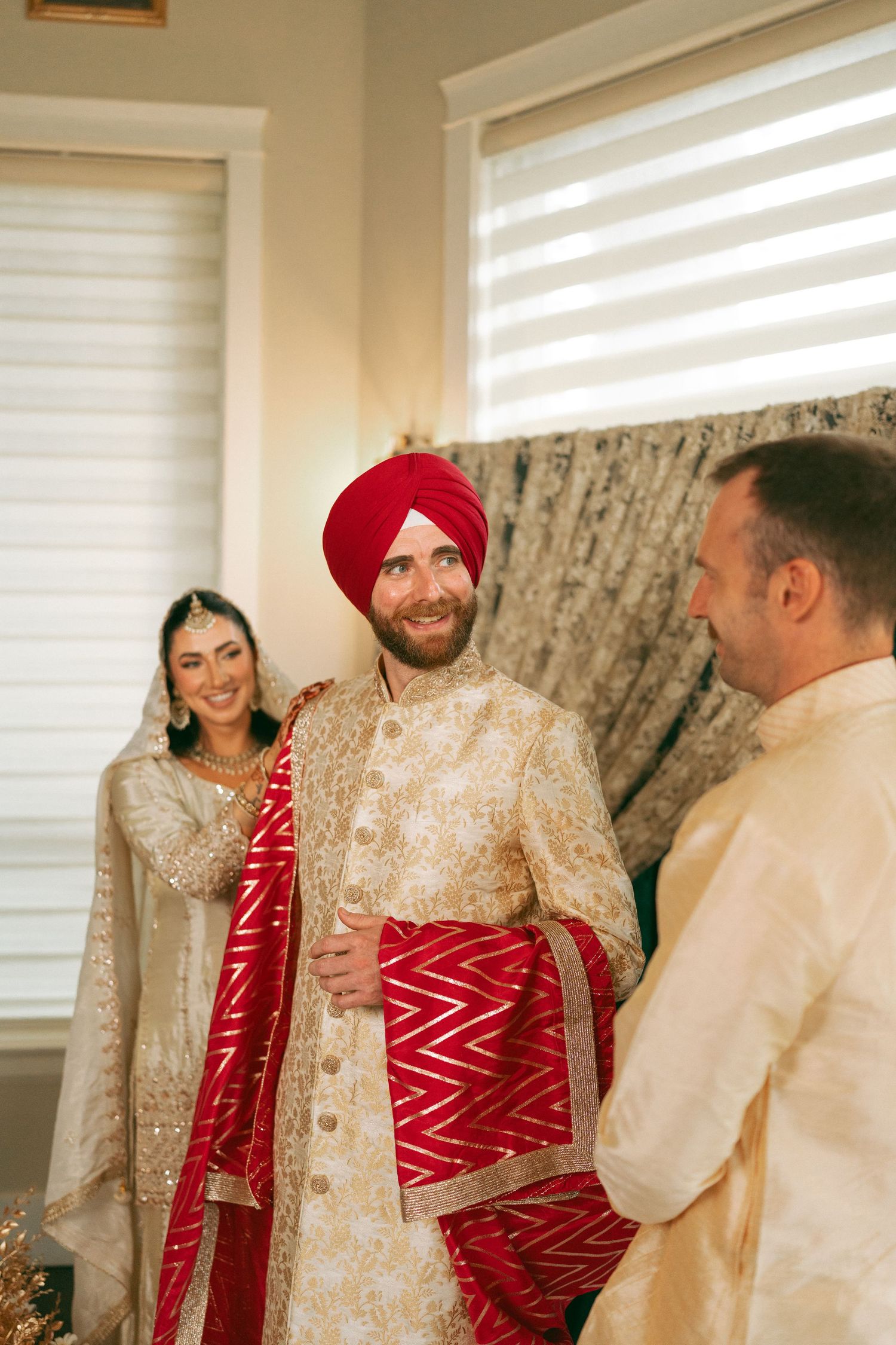 Sikh groom in red turban and gold sherwani smiles with bride in white and red lehenga during wedding ceremony.