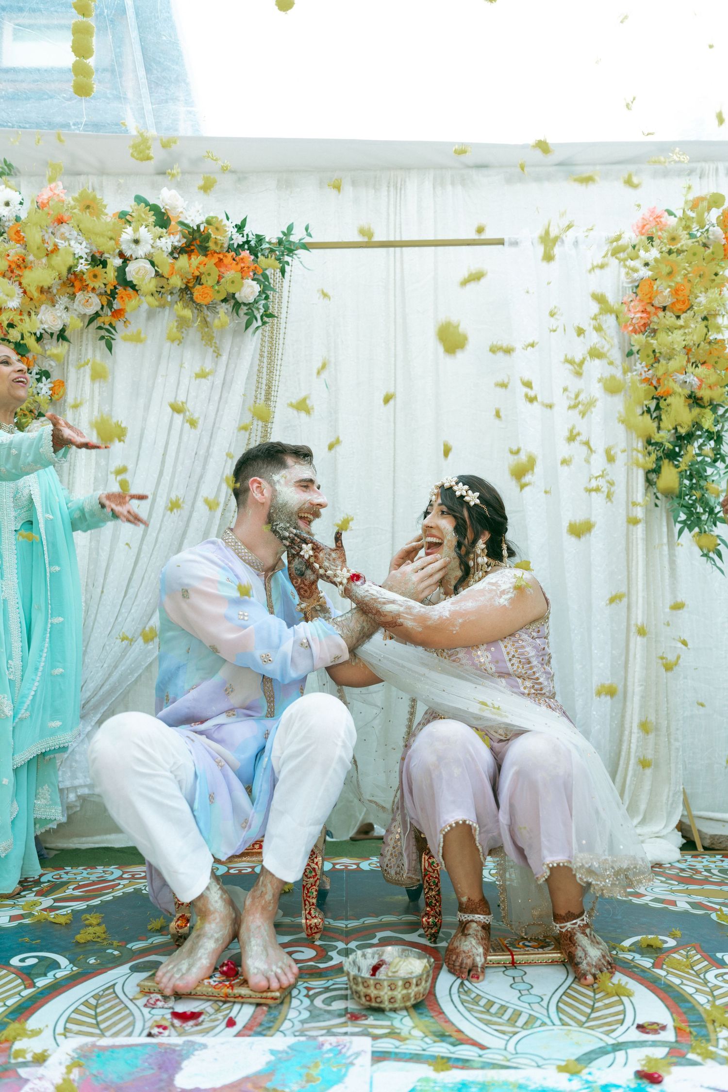 Couple laughing during Indian Haldi ceremony with flower petals falling, decorated with floral backdrop.