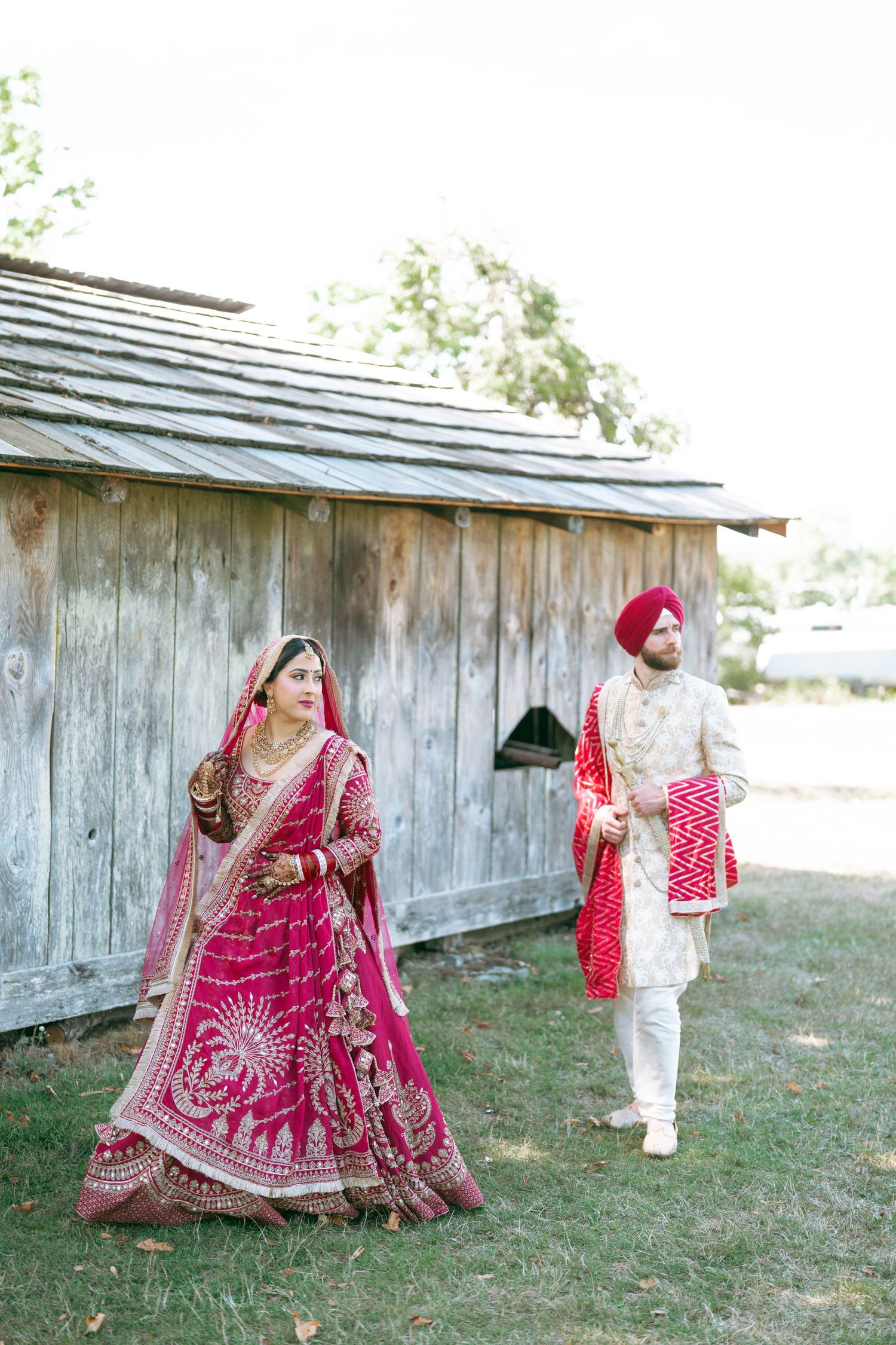 Indian bride in red lehenga and groom in white sherwani with turban stand outside rustic wooden barn.