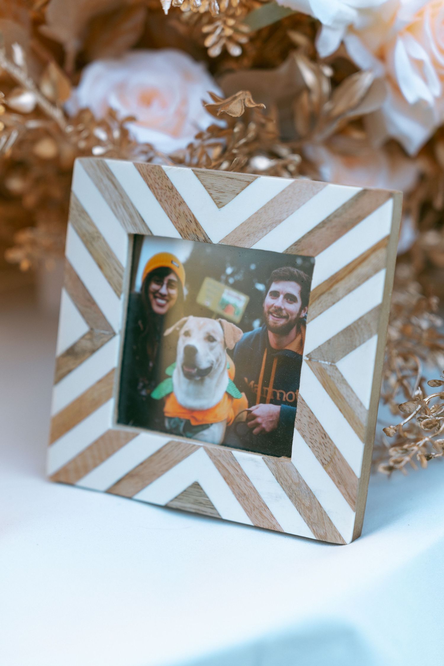 Decorative chevron wood picture frame with a photo of two people and a dog, surrounded by blush floral arrangement.