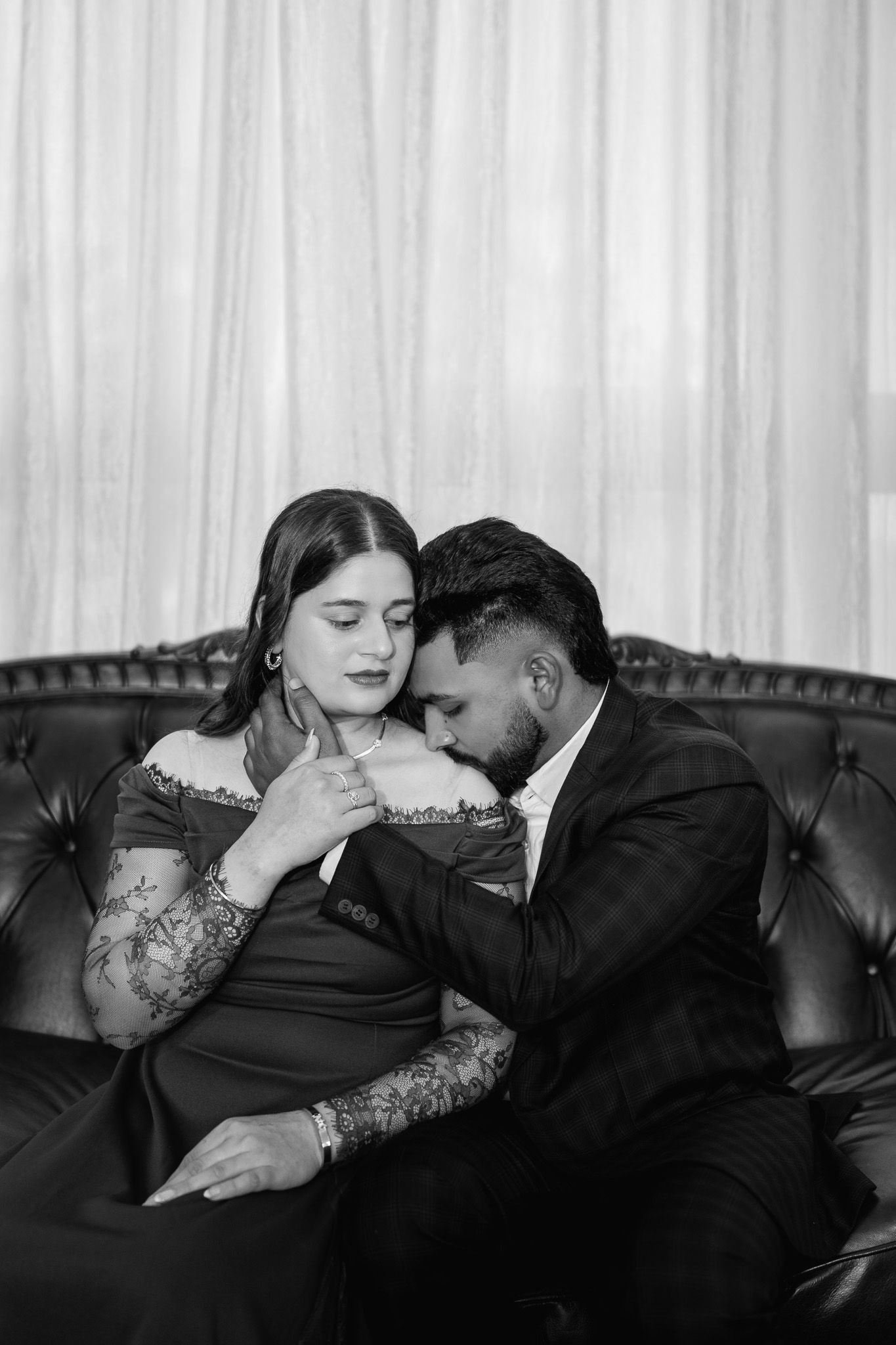 Black and white photo of a couple sharing an intimate moment on a tufted leather sofa, woman in elegant dress with tattoos.