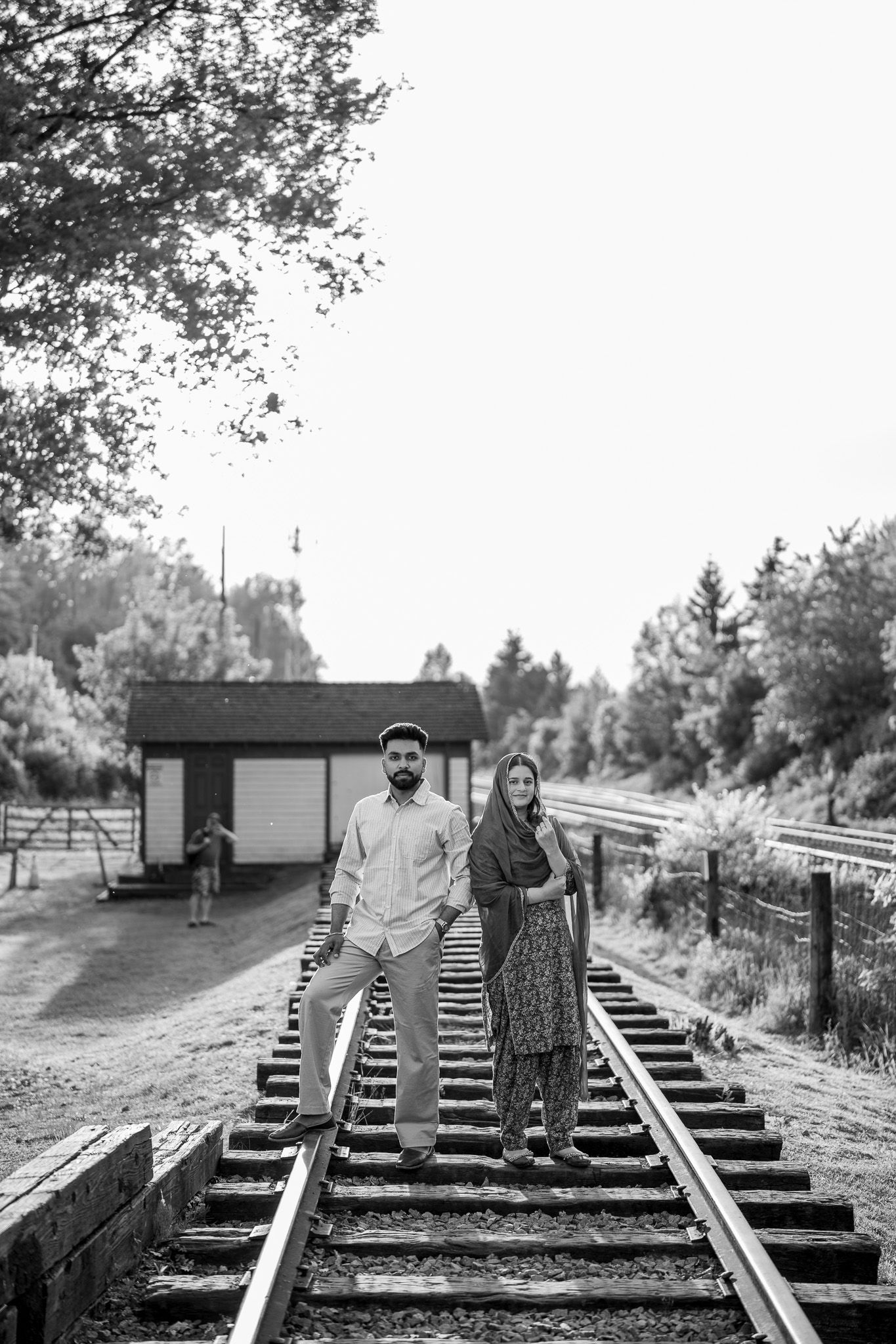 Black and white photo of a South Asian couple standing together on railway tracks surrounded by trees.