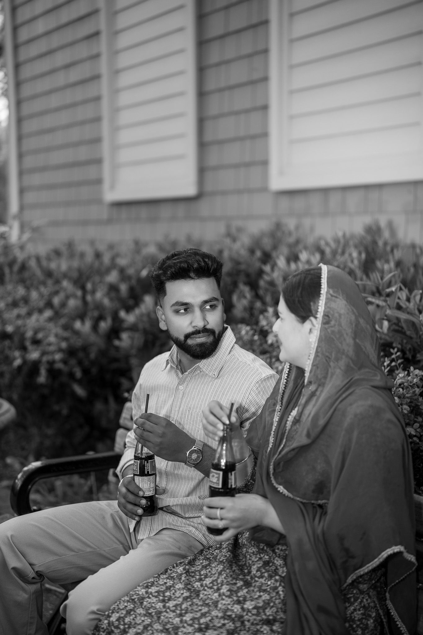 Black and white photo of couple sitting outdoors sharing drinks near a garden bush.