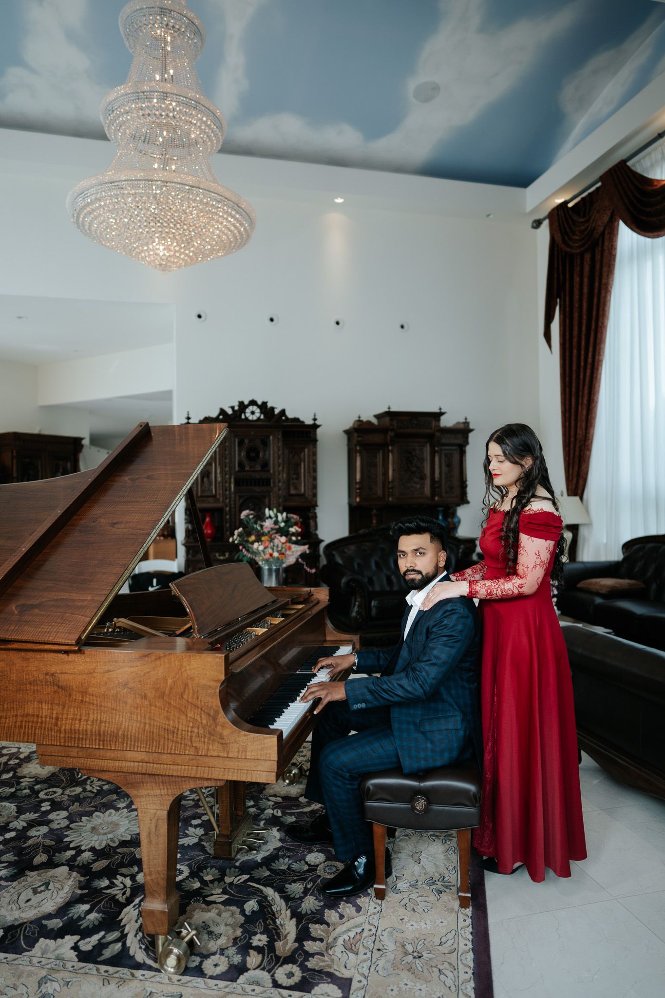 Couple in elegant attire poses by a grand piano in a luxurious room with crystal chandelier.