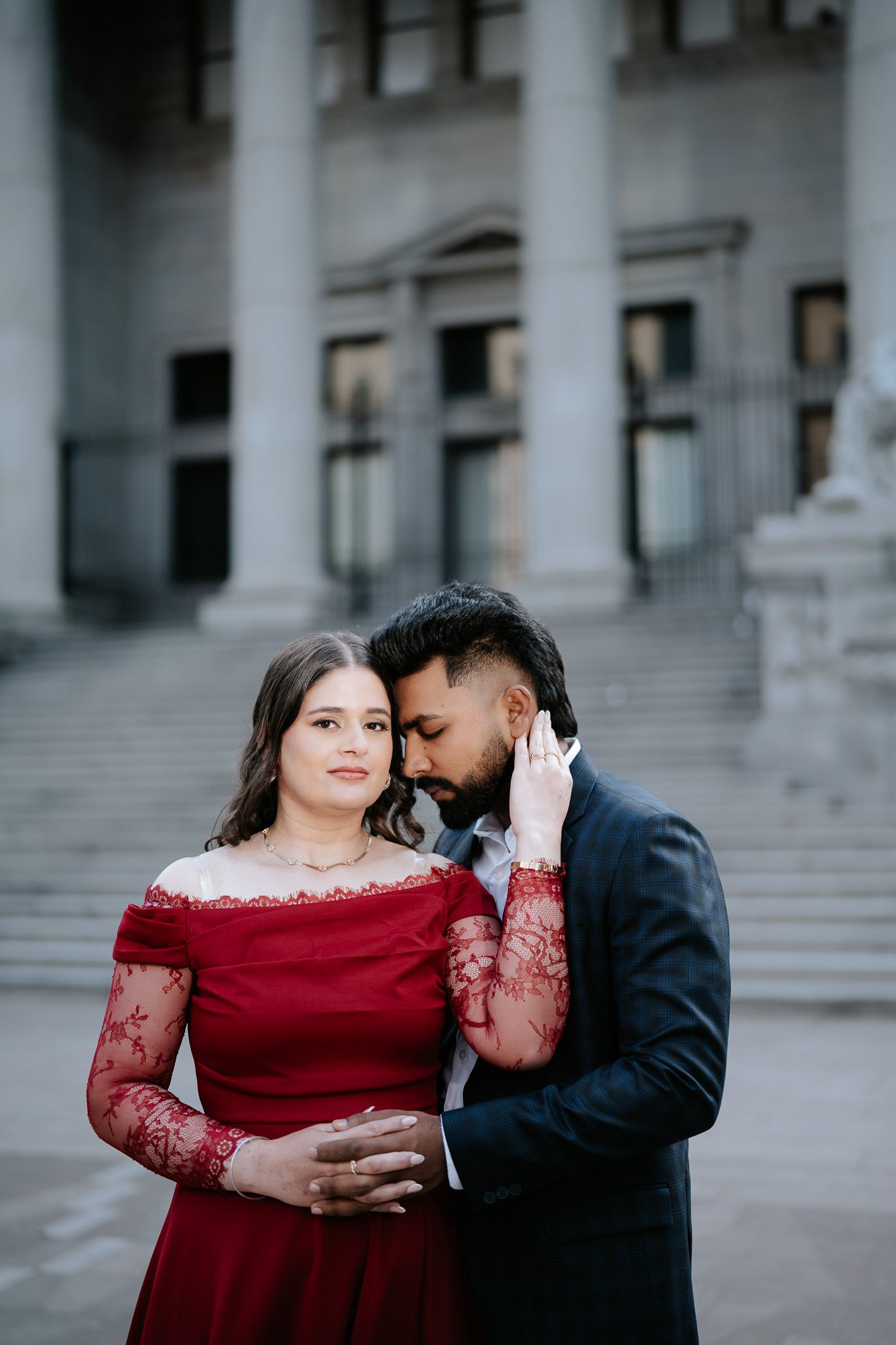 Couple in elegant attire embrace on grand steps, woman in red lace gown, man in dark suit whispering tenderly.