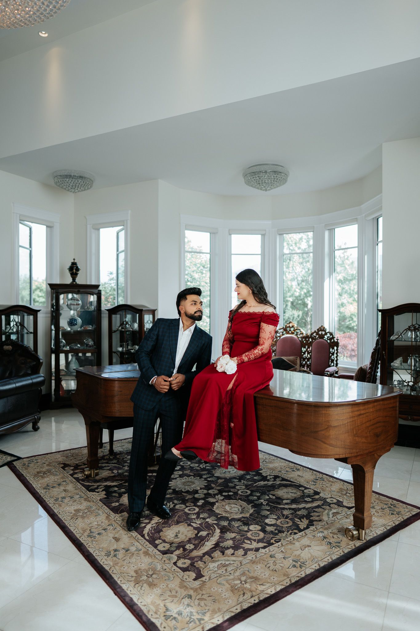 Elegant couple posing with grand piano in bright white room, woman in red gown holding white roses, man in dark suit.