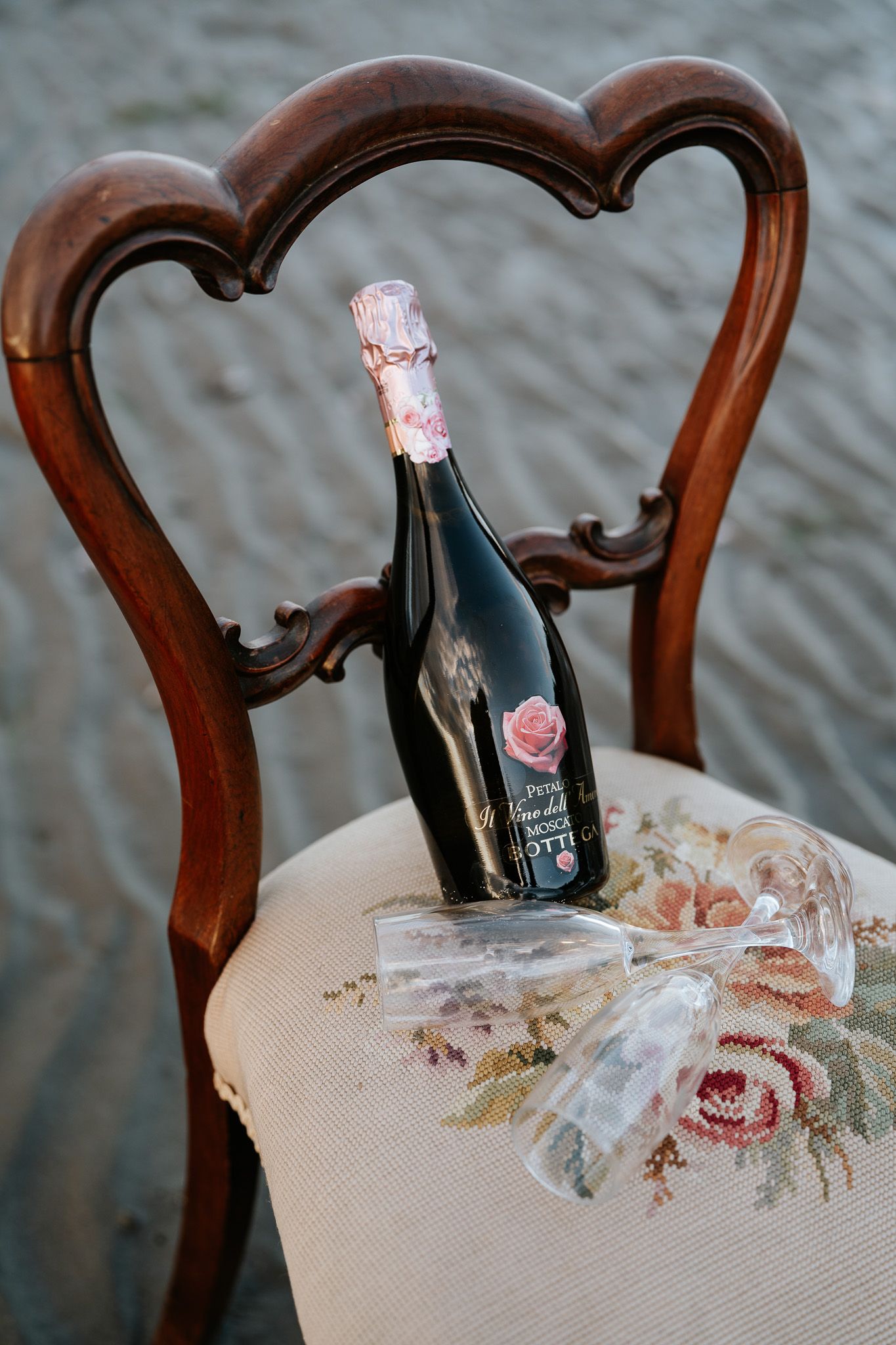 Elegant dark wine bottle with floral label resting on a vintage wooden chair with embroidered floral cushion.