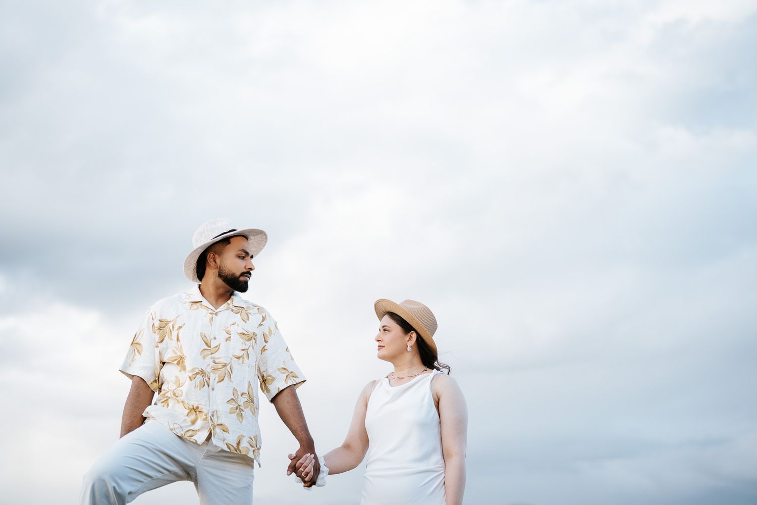 Interracial couple holding hands in matching white outfits and hats against a cloudy sky.