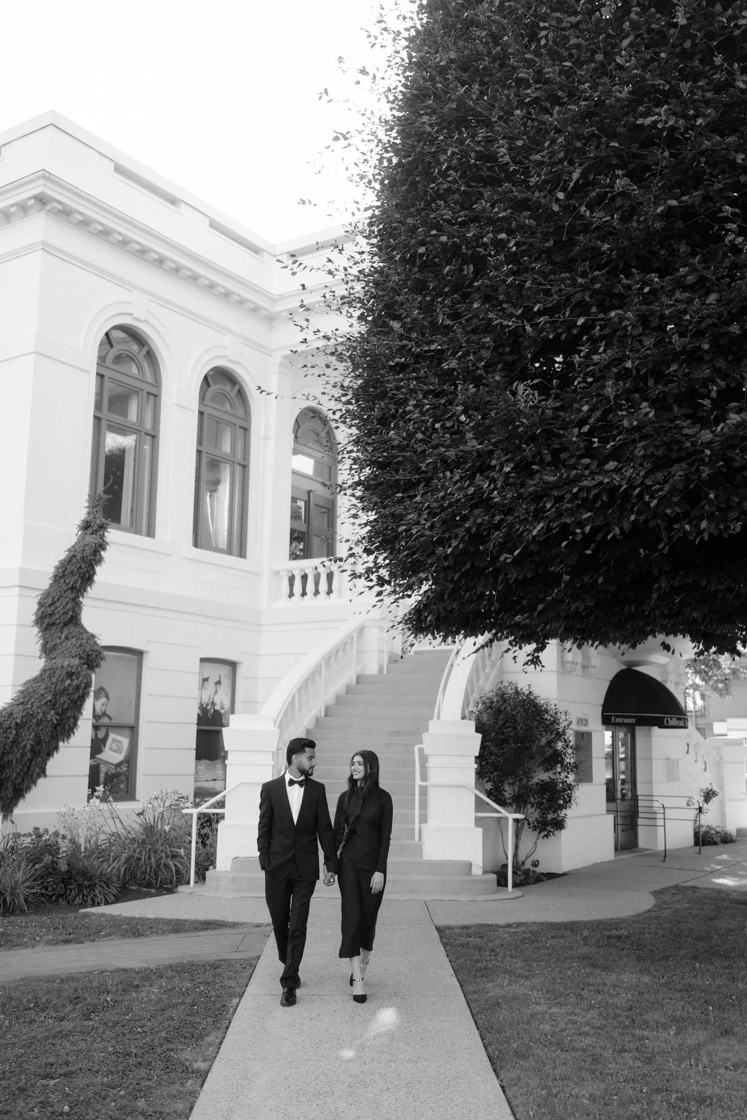 Elegant couple in matching black outfits walking hand in hand outside a grand white building, black and white photo.