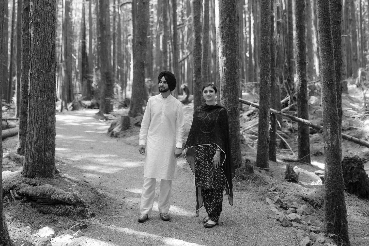A Sikh couple walks hand in hand along a forest path, dressed in traditional attire, in a black and white photo.