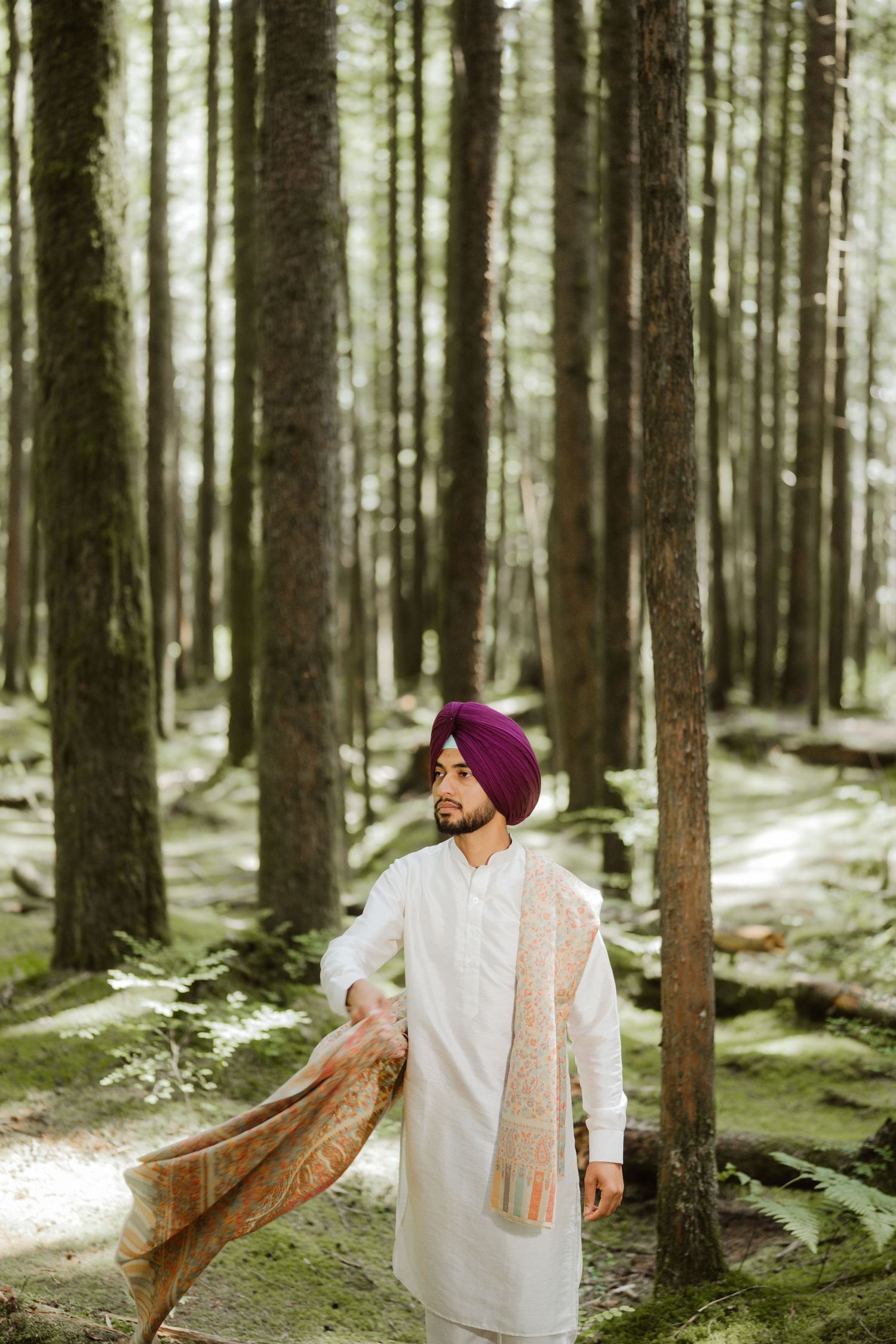 Sikh man in white kurta and purple turban holding a decorative shawl while standing among tall trees in a forest.