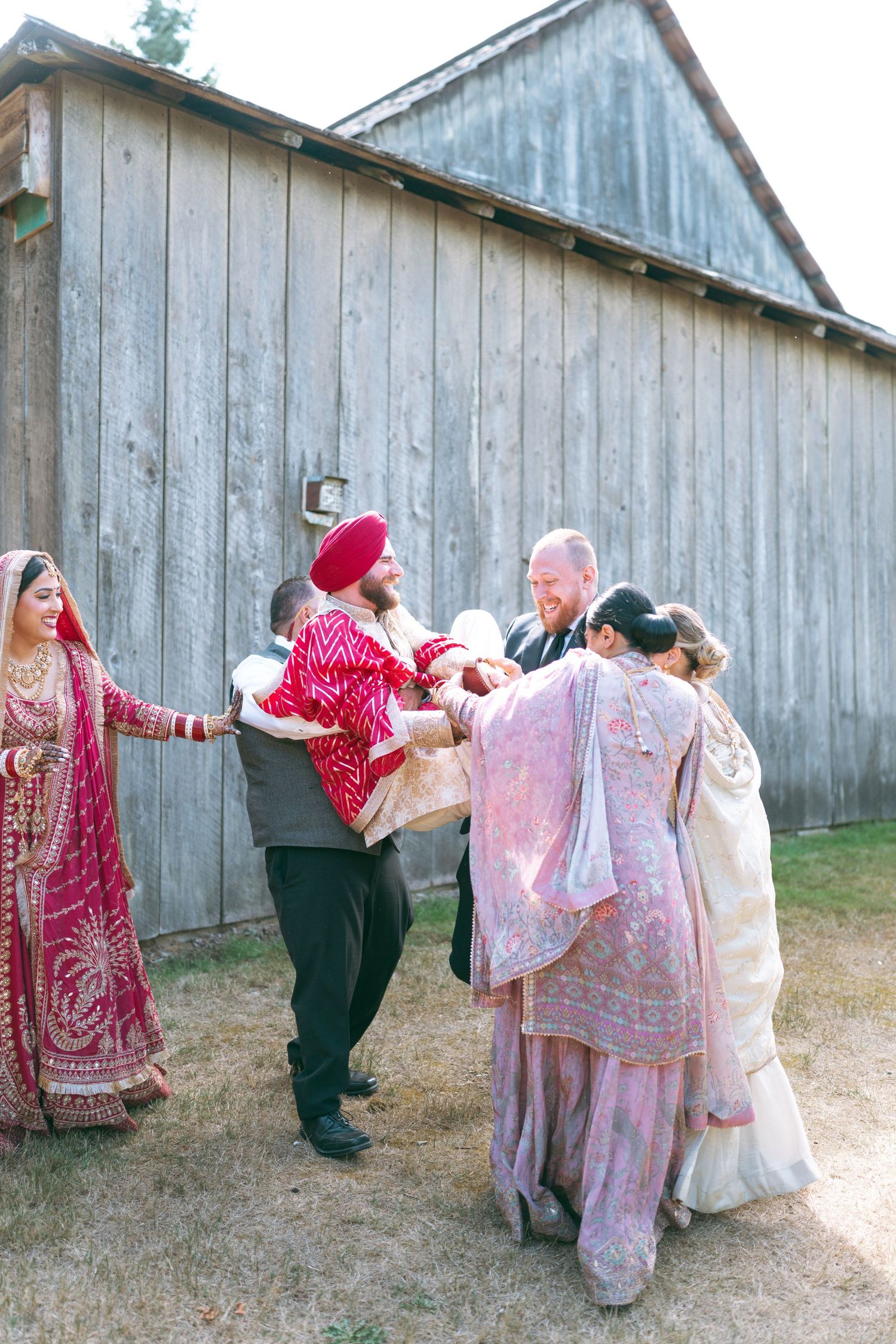 South Asian wedding party dancing joyfully outside a rustic barn, bride in red lehenga and groom in turban.
