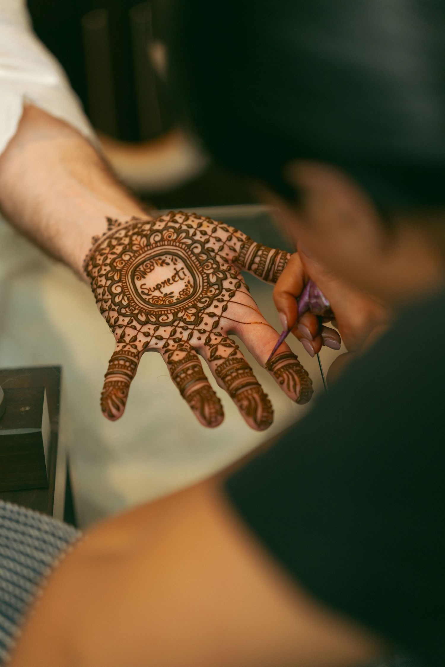 Artist applying intricate henna mehndi design on a palm, featuring geometric patterns and text centerpiece.