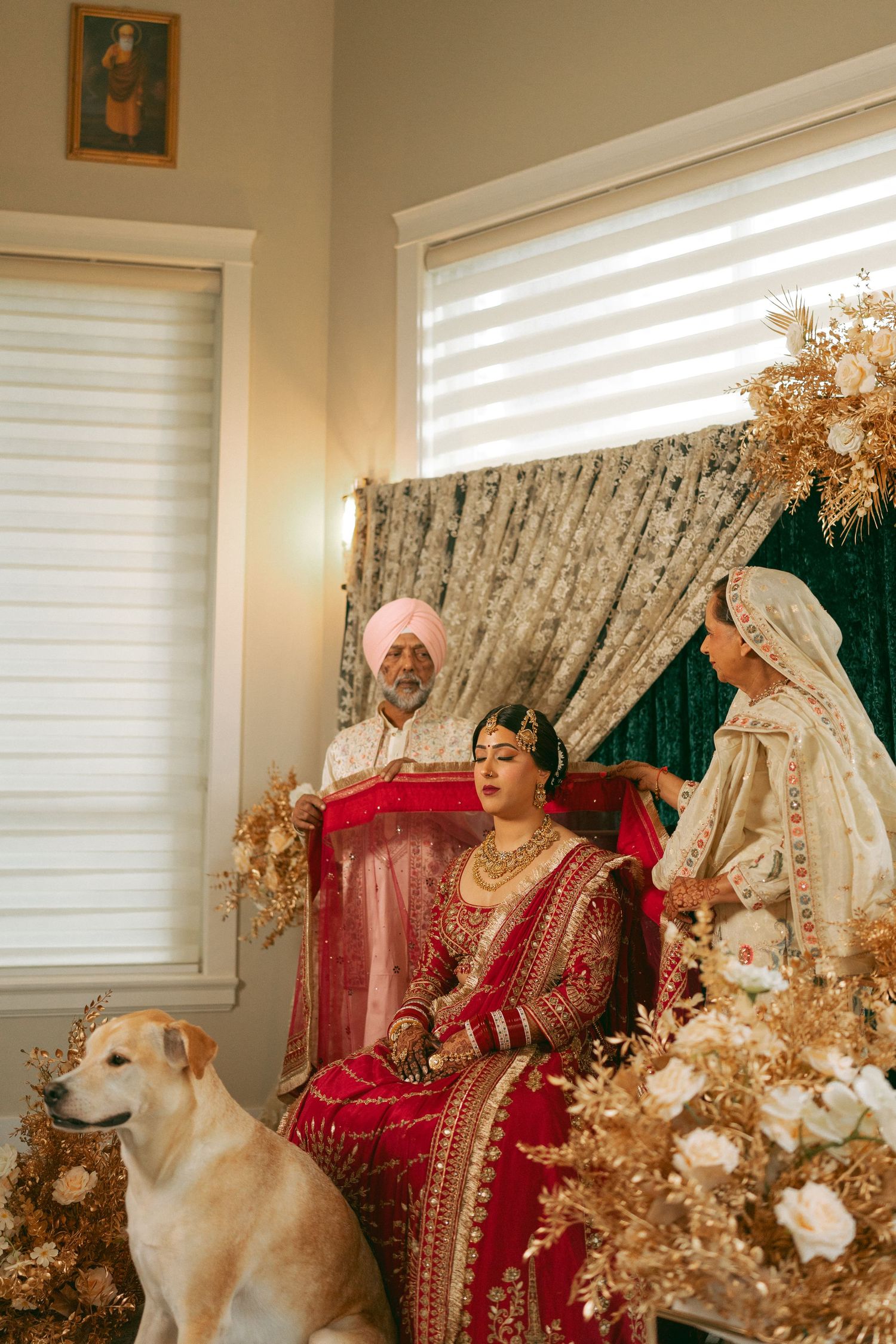 Sikh bride in red lehenga seated at anand karaj ceremony with family and a white dog amid golden floral decor.