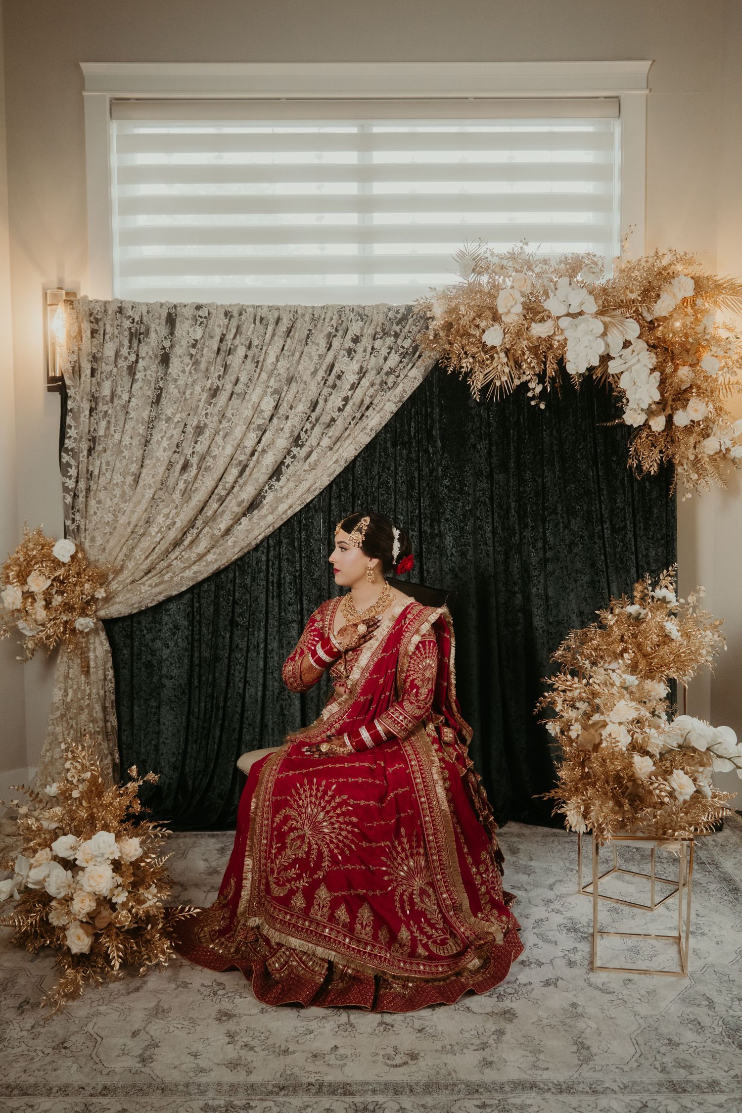 Bride in red and gold lehenga sits elegantly before dark floral backdrop with dried flowers and lace curtains.
