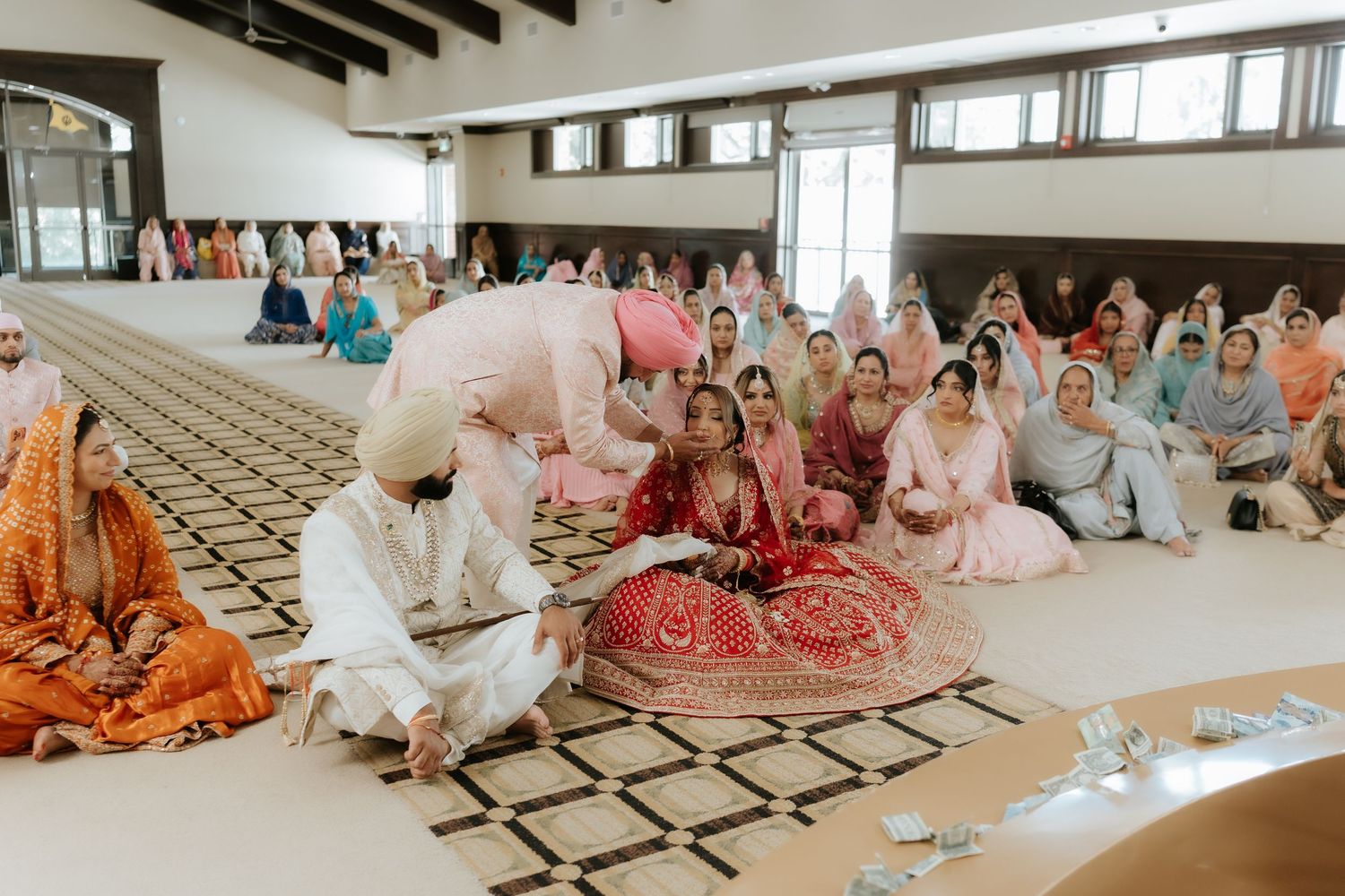 Sikh wedding ceremony with bride in red lehenga surrounded by guests in colorful attire inside a gurudwara.