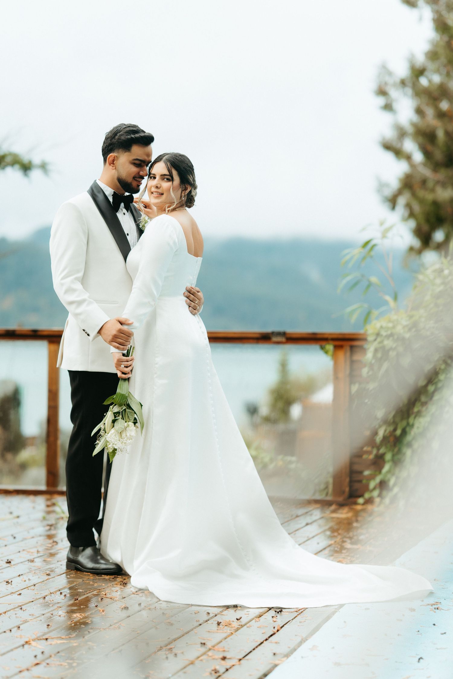 A bride and groom share an intimate moment on a wooden deck overlooking a scenic mountain landscape.