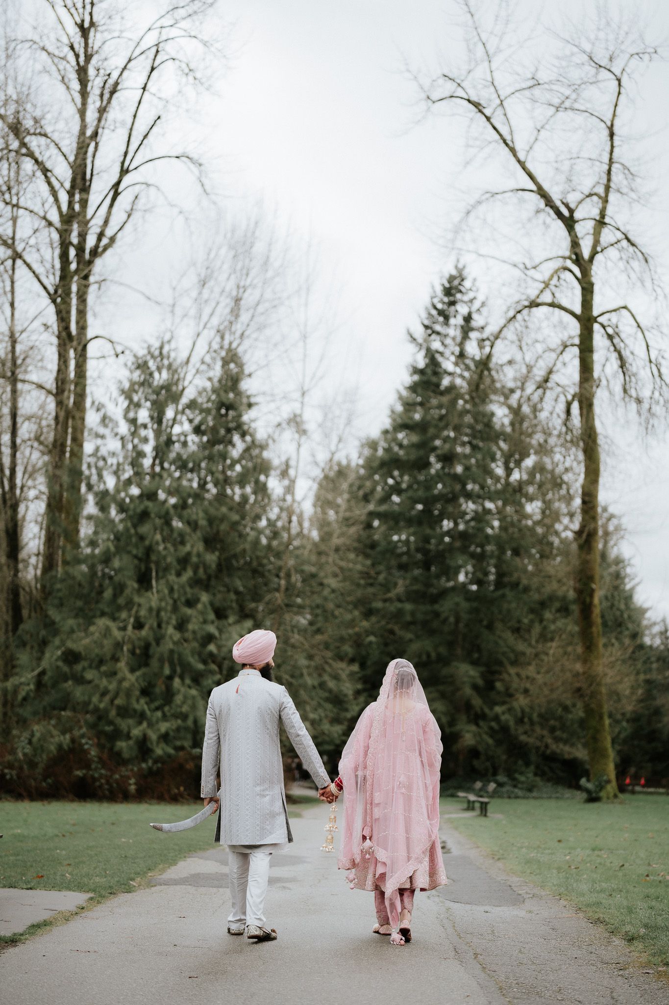 A couple in traditional attire walks along a tree-lined path in a serene park setting during winter.