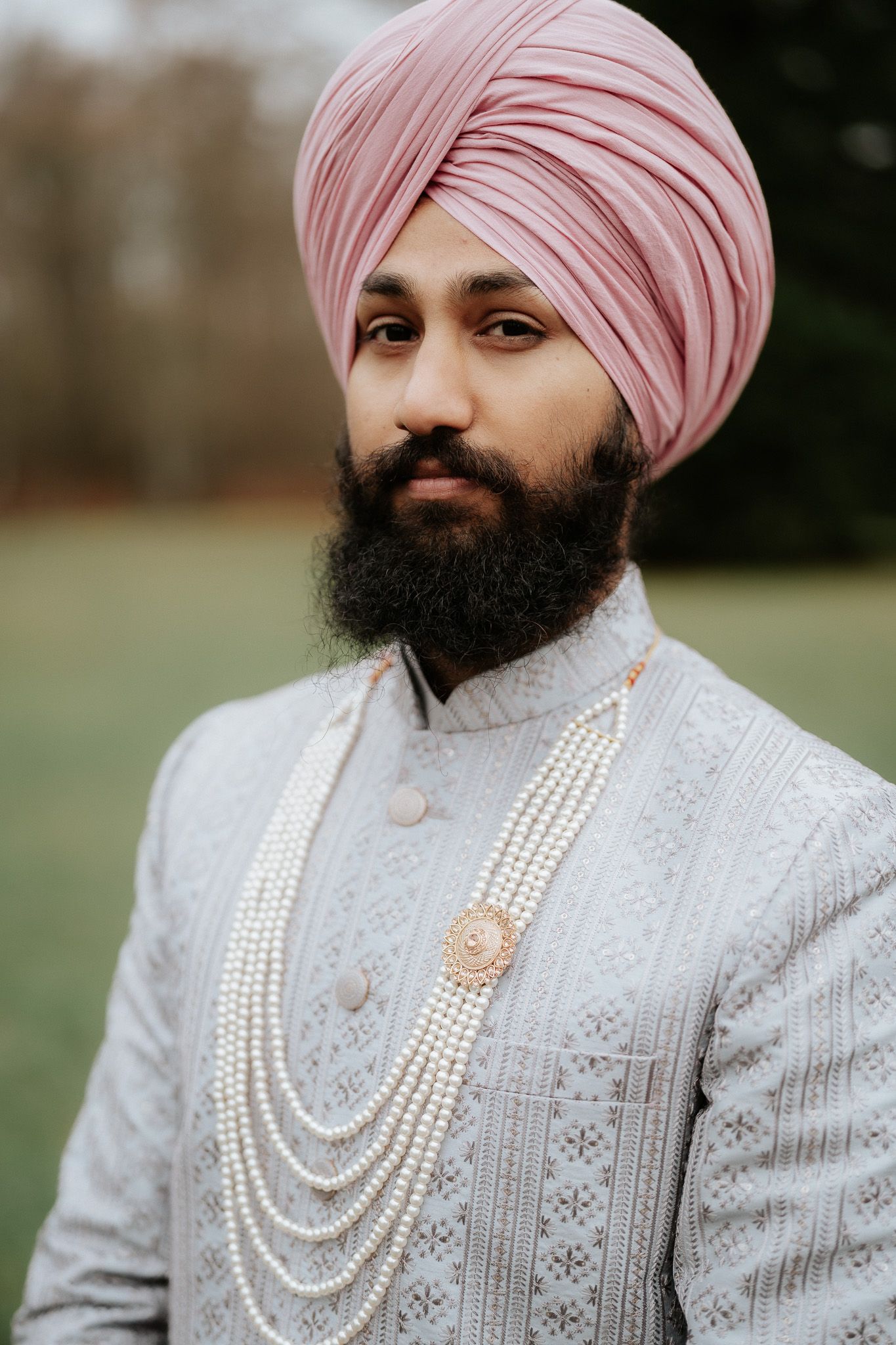 A groom in a traditional Sikh wedding attire wearing an embroidered white sherwani, pink turban and layered pearl necklaces.