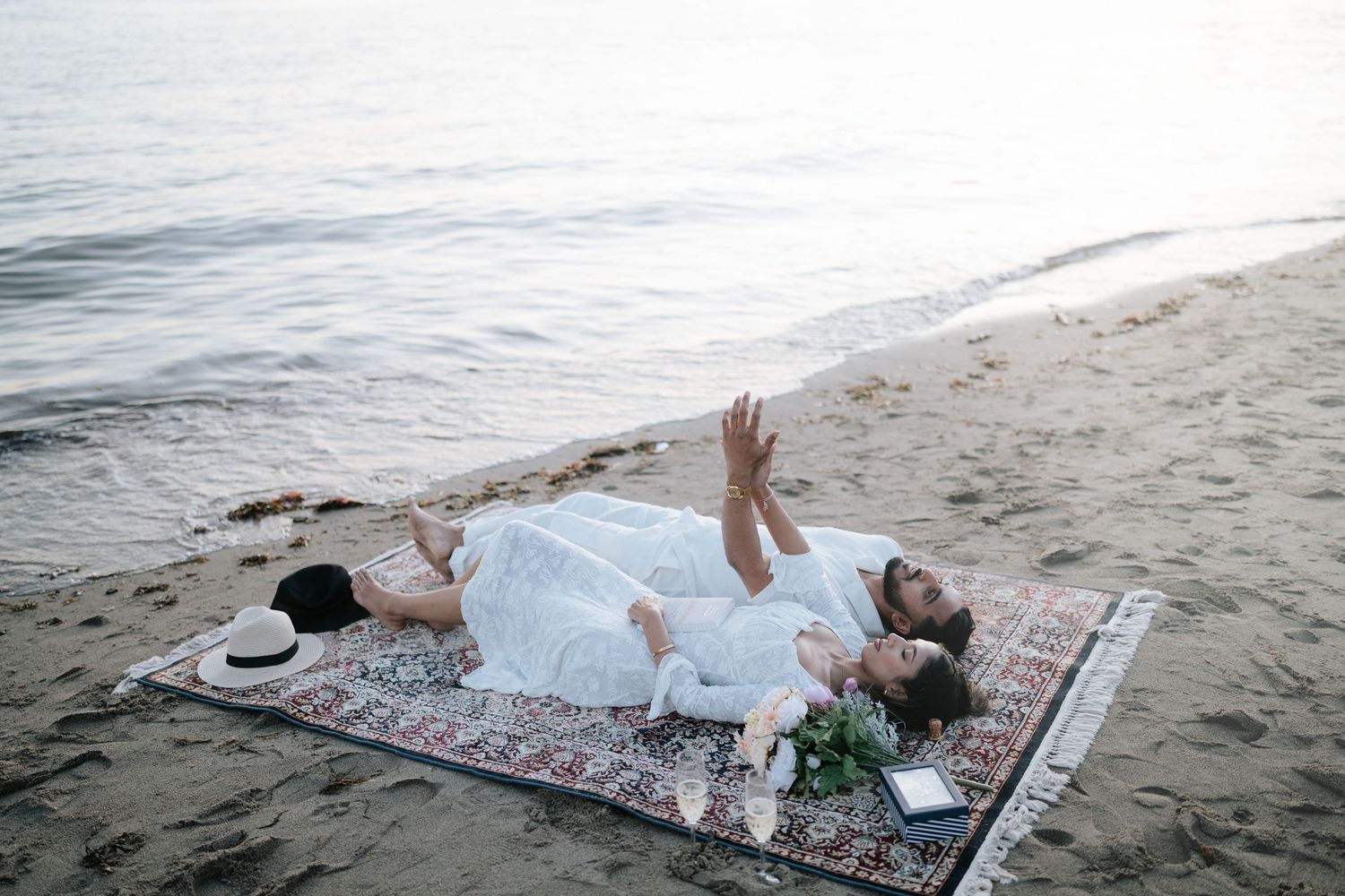 Two people relax on a patterned blanket at the beach while waves gently roll onto the shore.