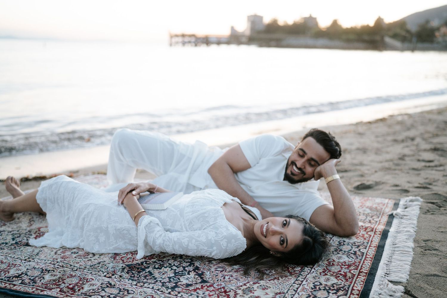 A person in a white dress lies gracefully on a patterned rug at the beach during sunset.