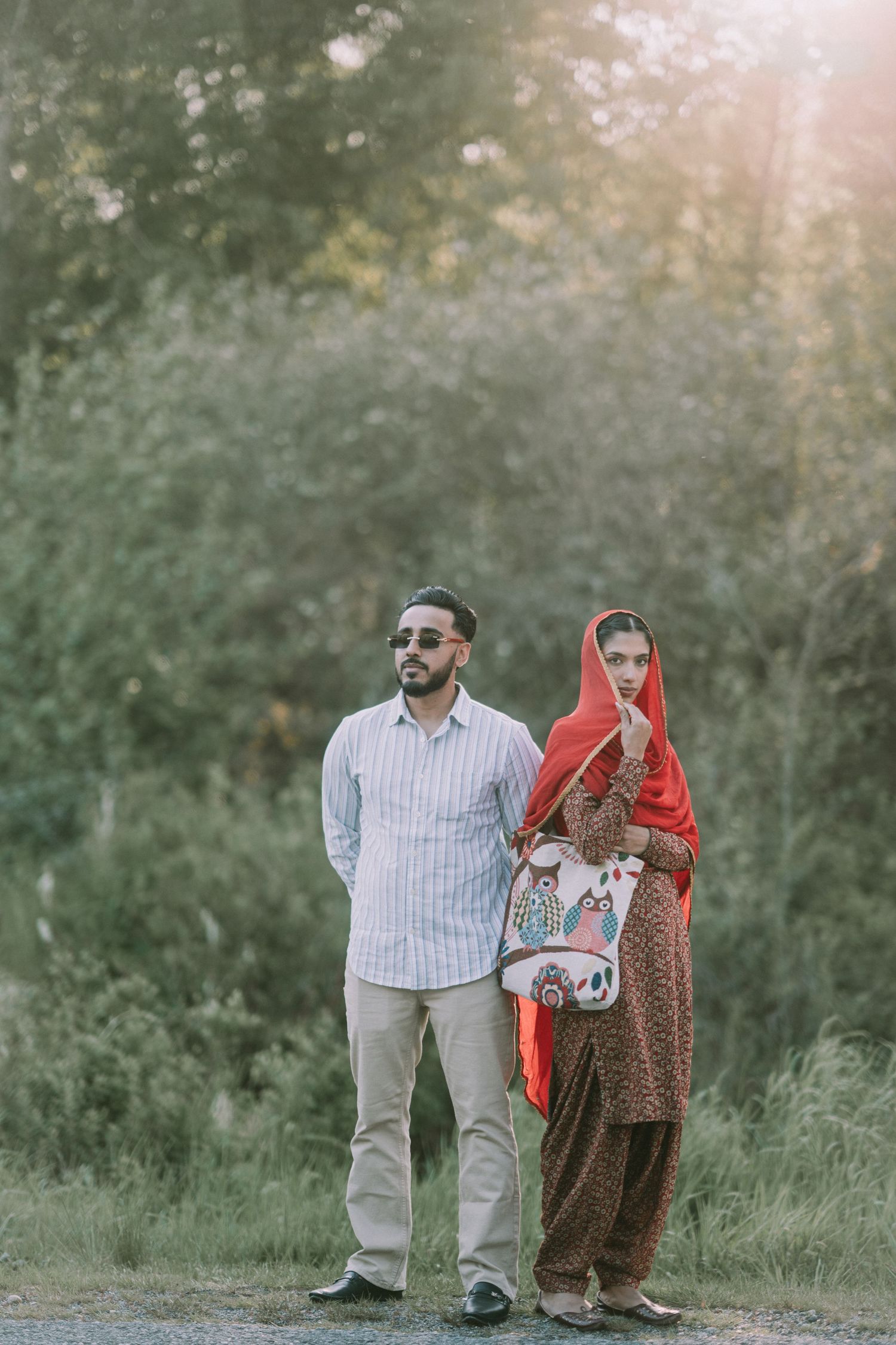 A couple in traditional Punjabi attire poses playfully in a grassy outdoor setting during golden hour.