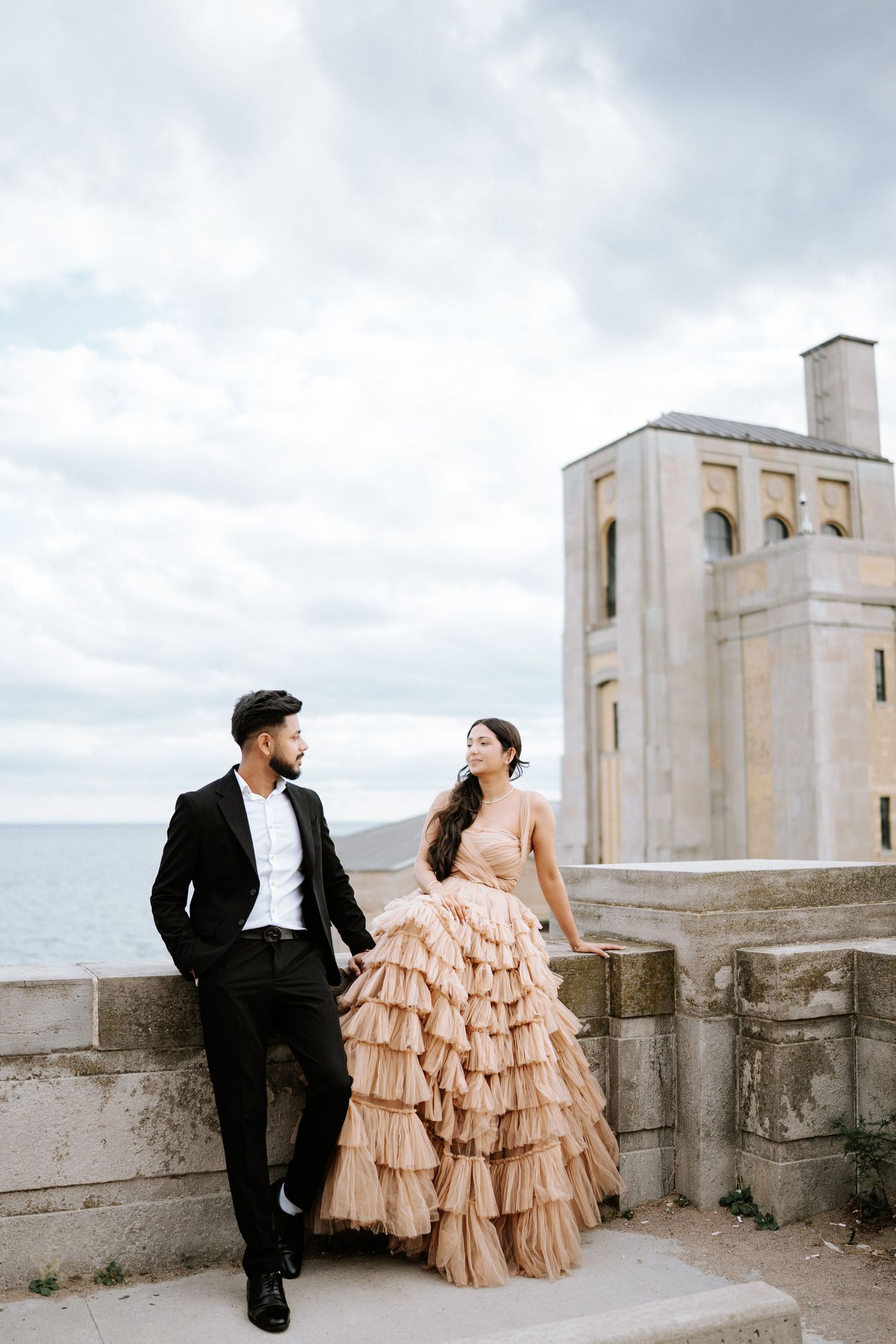 Romantic couple in formal wear posing against architectural backdrop with cloudy sky.