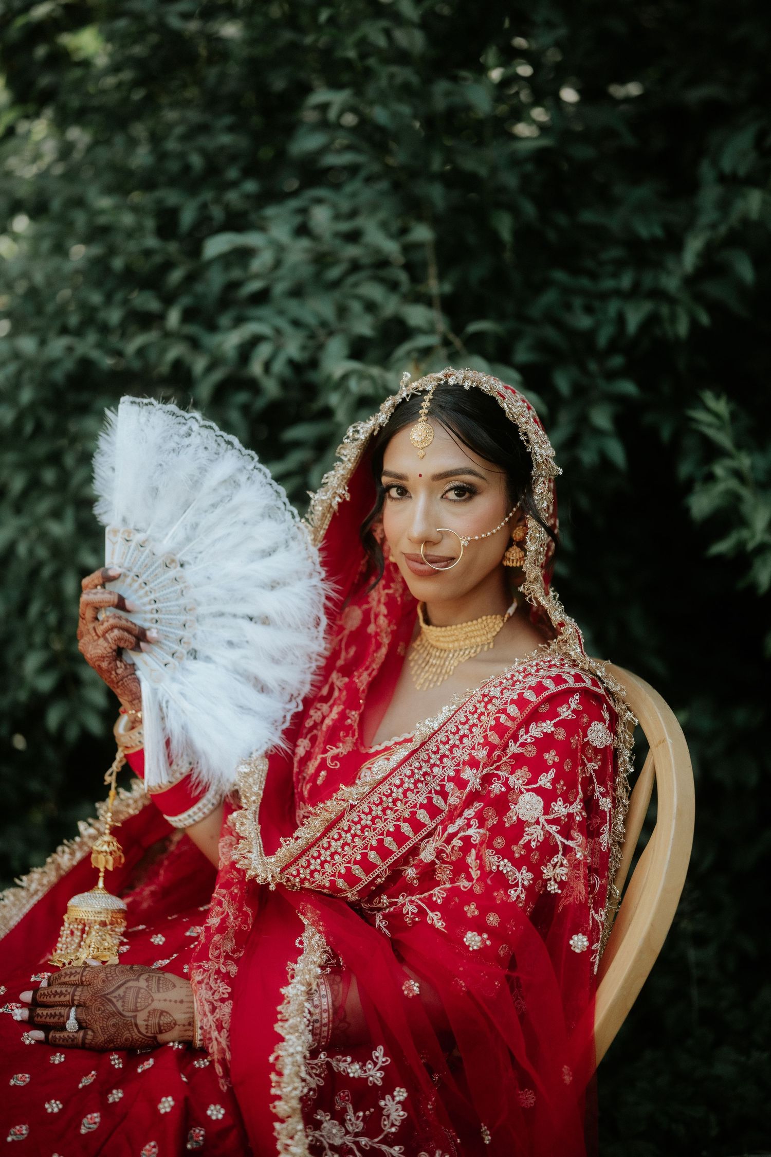 A bride in a red embroidered lehenga poses with an elegant white fan against a dark green background.
