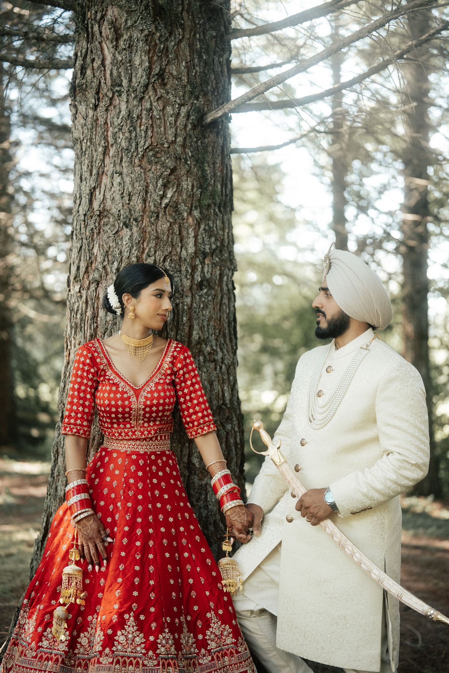 A couple in red and white traditional Indian wedding outfits pose in a forest setting.