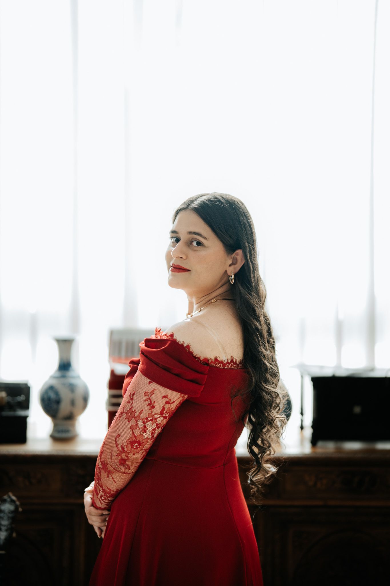 Young woman in red off-shoulder lace gown poses gracefully in front of sheer curtains and antique furniture.