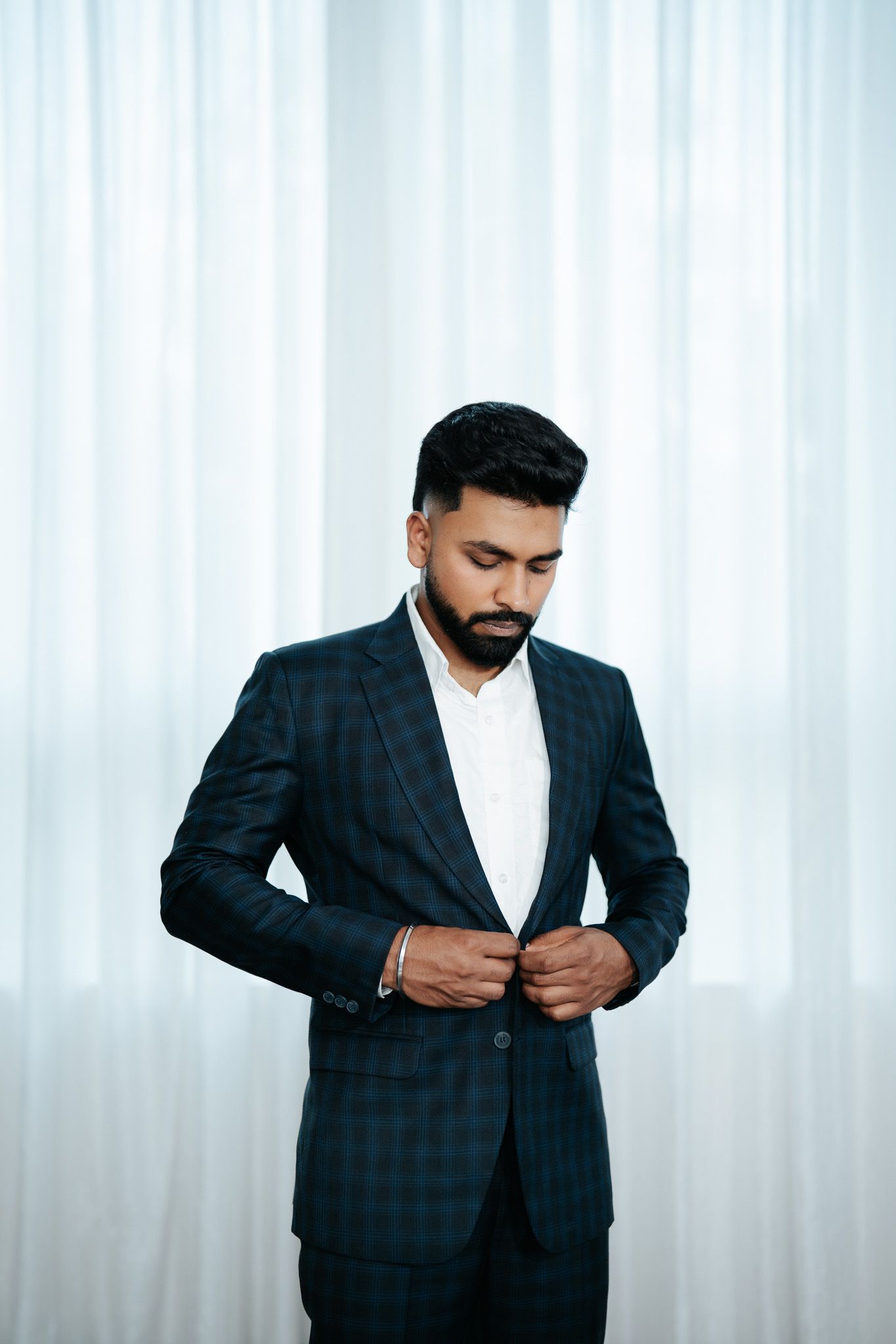 Stylish bearded man in dark plaid suit looking down while adjusting jacket buttons against white curtains.