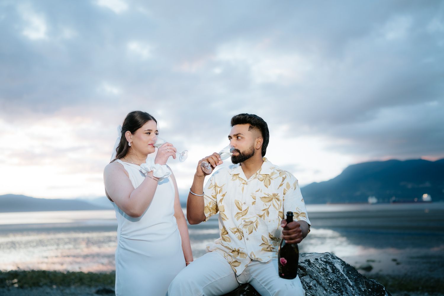 Couple sharing a romantic toast with champagne by a scenic mountain lake at sunset, dressed in elegant white outfits.