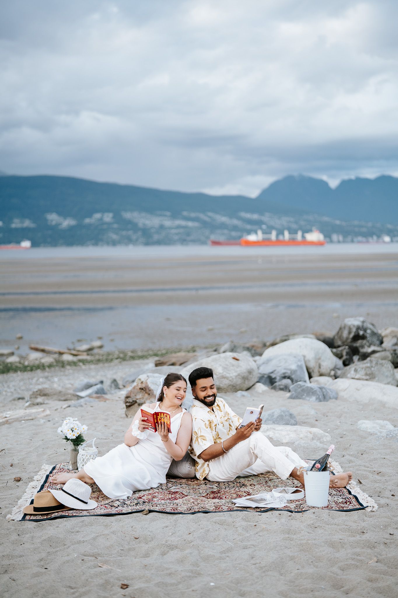 Couple sitting on beach blanket with drinks, mountains and ocean in background on cloudy day.